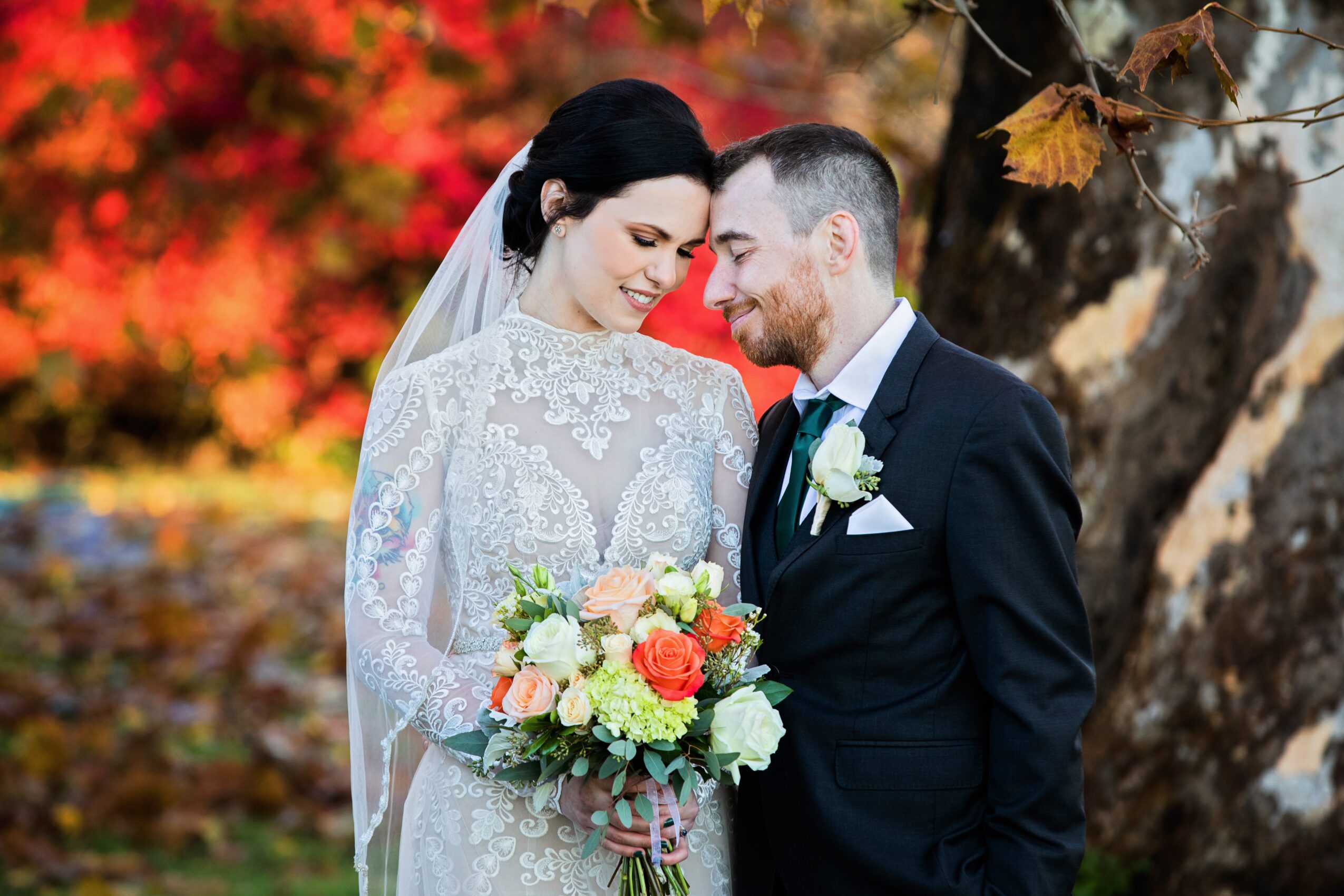 Bride and groom smiling close together outdoors, bride holding a bouquet, autumn leaves in background.