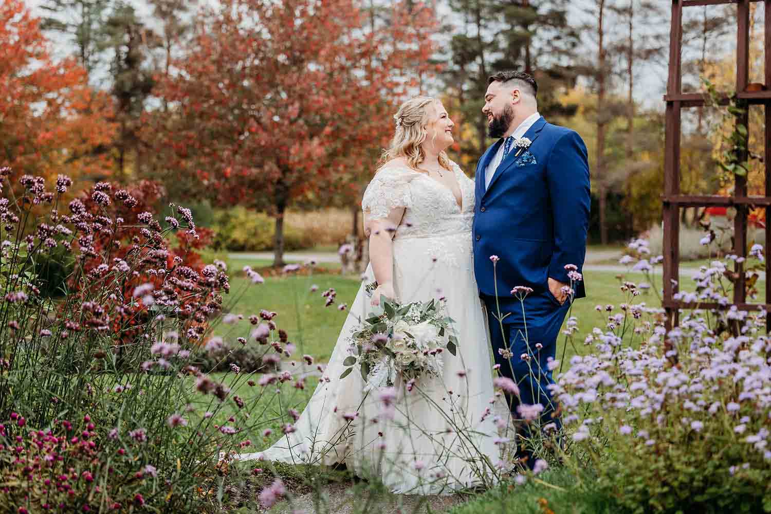 A bride and groom stand outdoors among colorful autumn trees and flowers, facing each other, holding hands, in wedding attire.