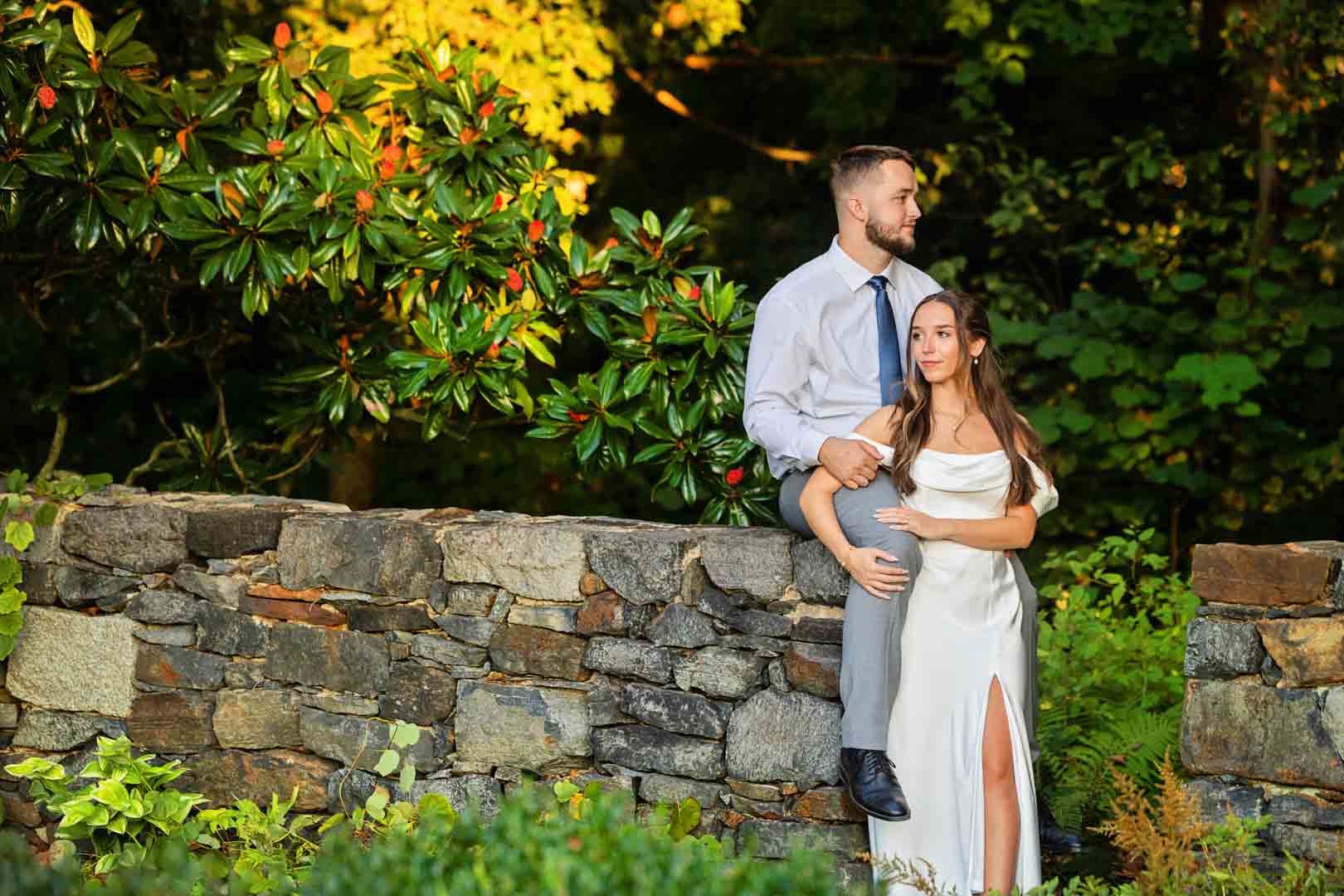 A couple stands on a stone wall in a garden with green foliage and yellow flowers, the woman in a white dress and the man in a light shirt and dark pants.