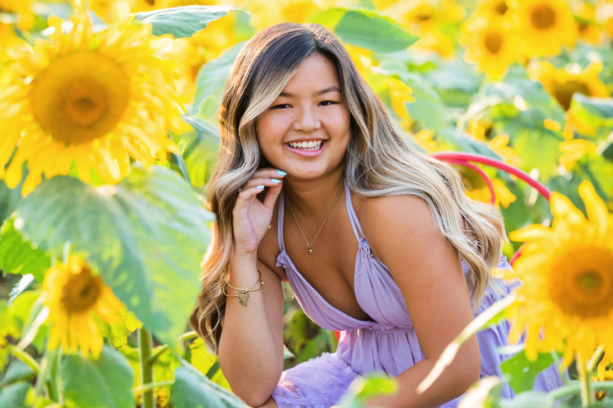 Smiling woman with long hair in a purple dress among sunflowers outdoors.