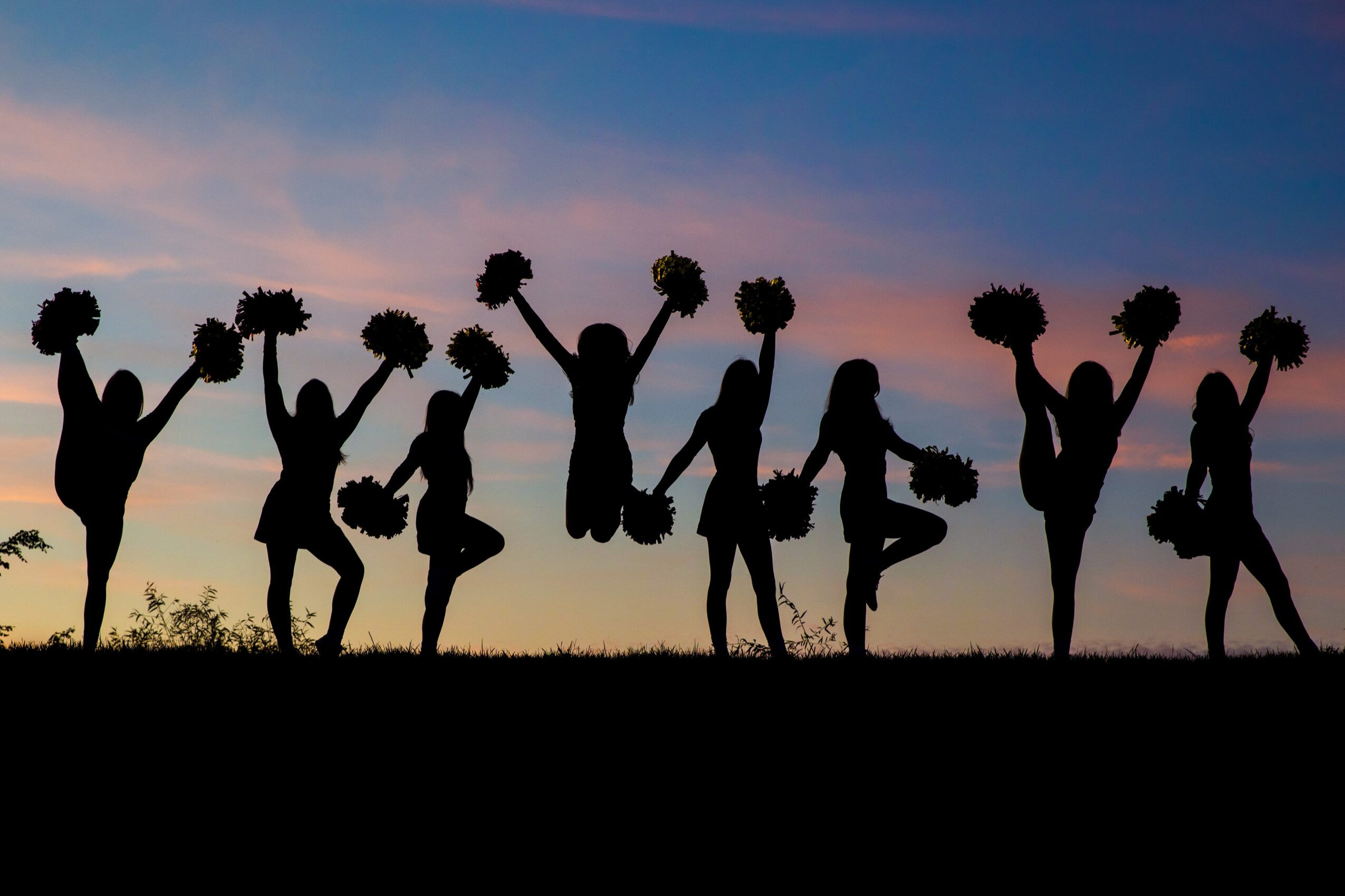 Silhouettes of children holding pom-poms and jumping against a sunset sky, celebrating outdoors.