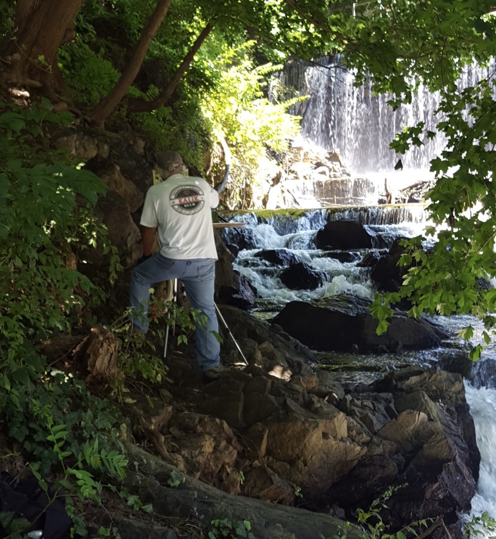 Person standing on rocks near a waterfall surrounded by green trees and foliage.