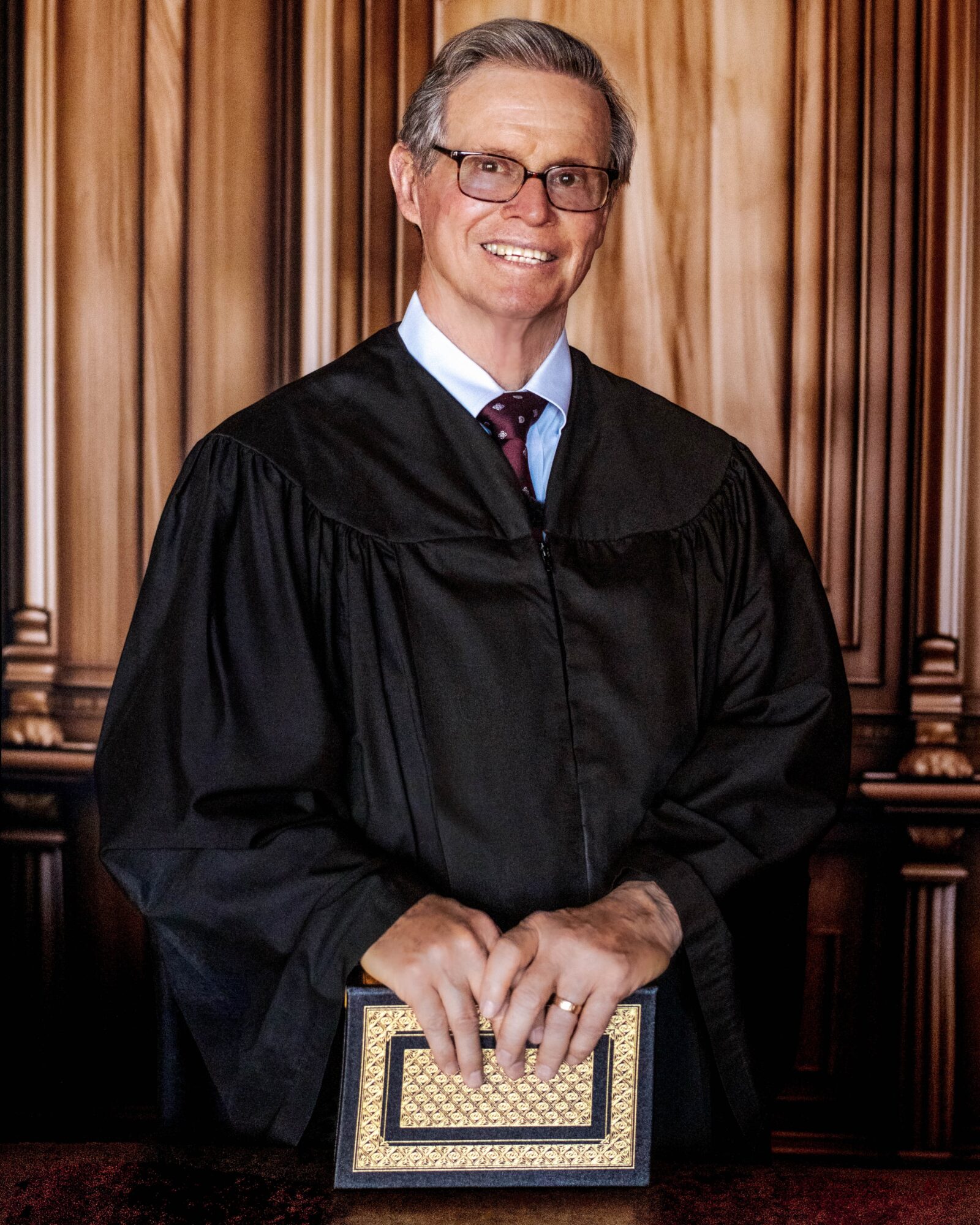 Man in a black gown holding a framed certificate, standing in front of wooden paneling, smiling, wearing glasses and a tie.