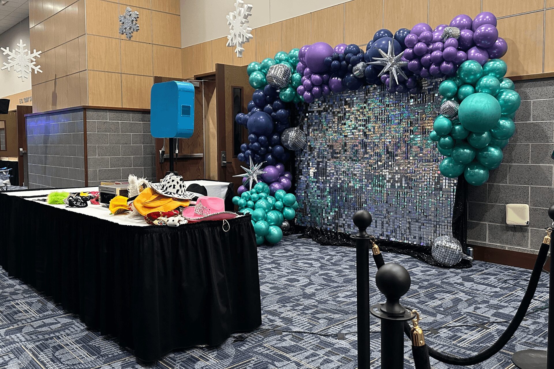 Balloon arch with purple, teal, and silver balloons, backdrop with glittering surface, and a table with party items in a decorated room.