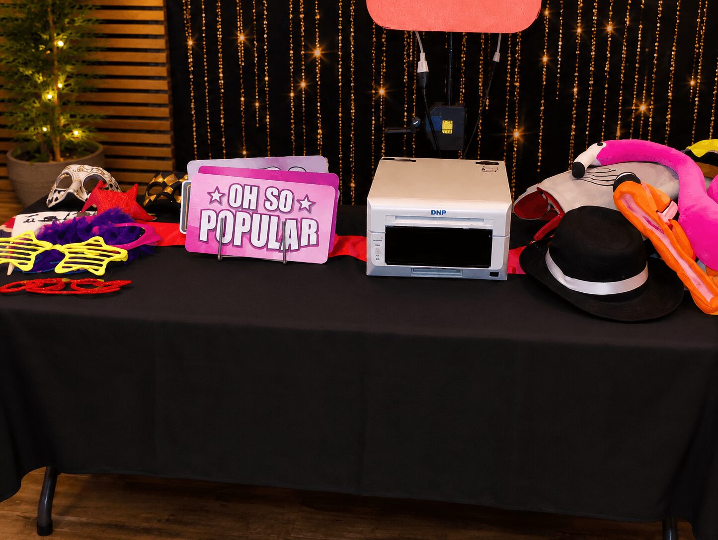 Table with party props, a pink sign reading 'OH SO POPULAR,' a microwave, and various accessories, against a decorated backdrop.