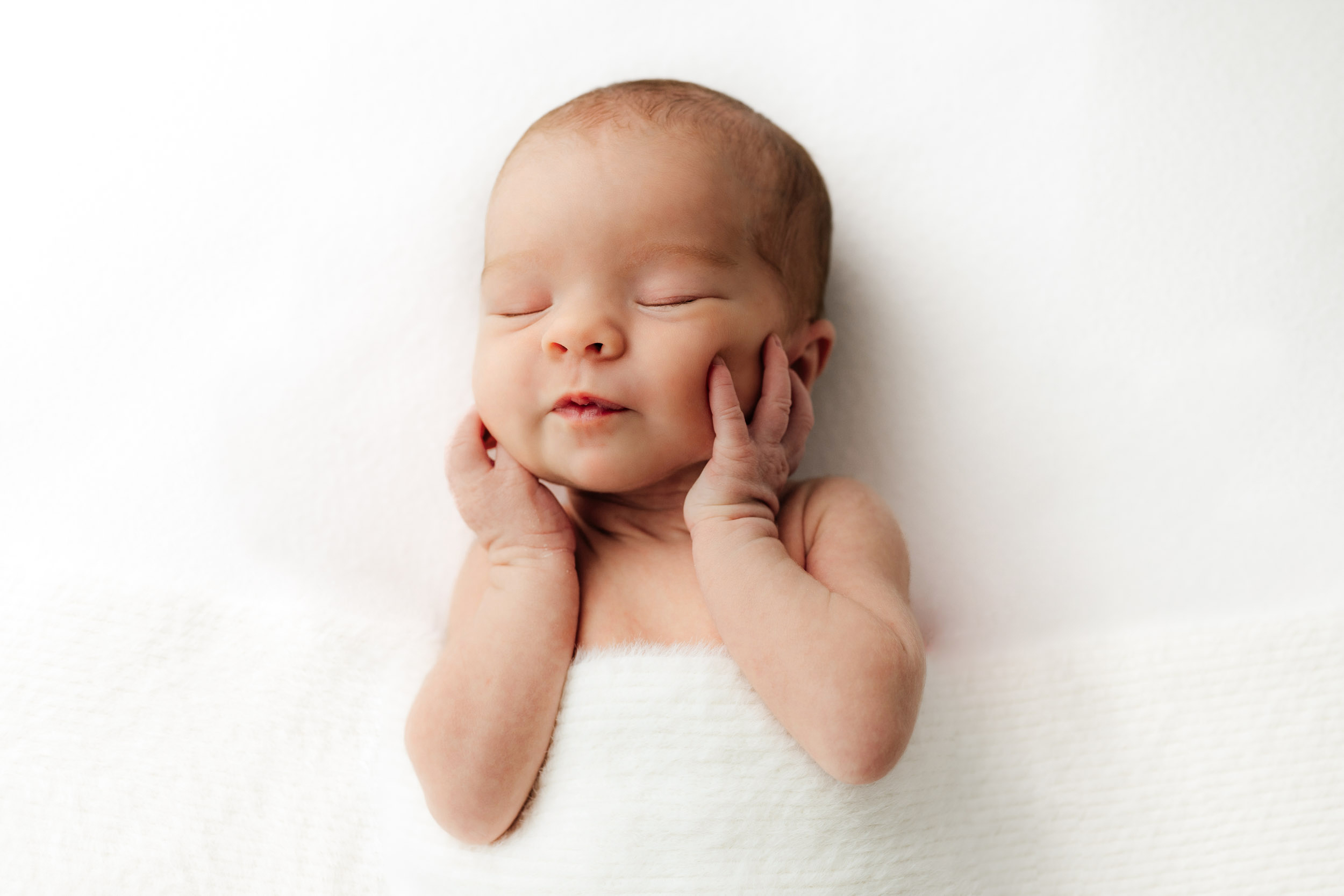 Baby with closed eyes, resting head on hands, wrapped in white cloth, lying on white background.