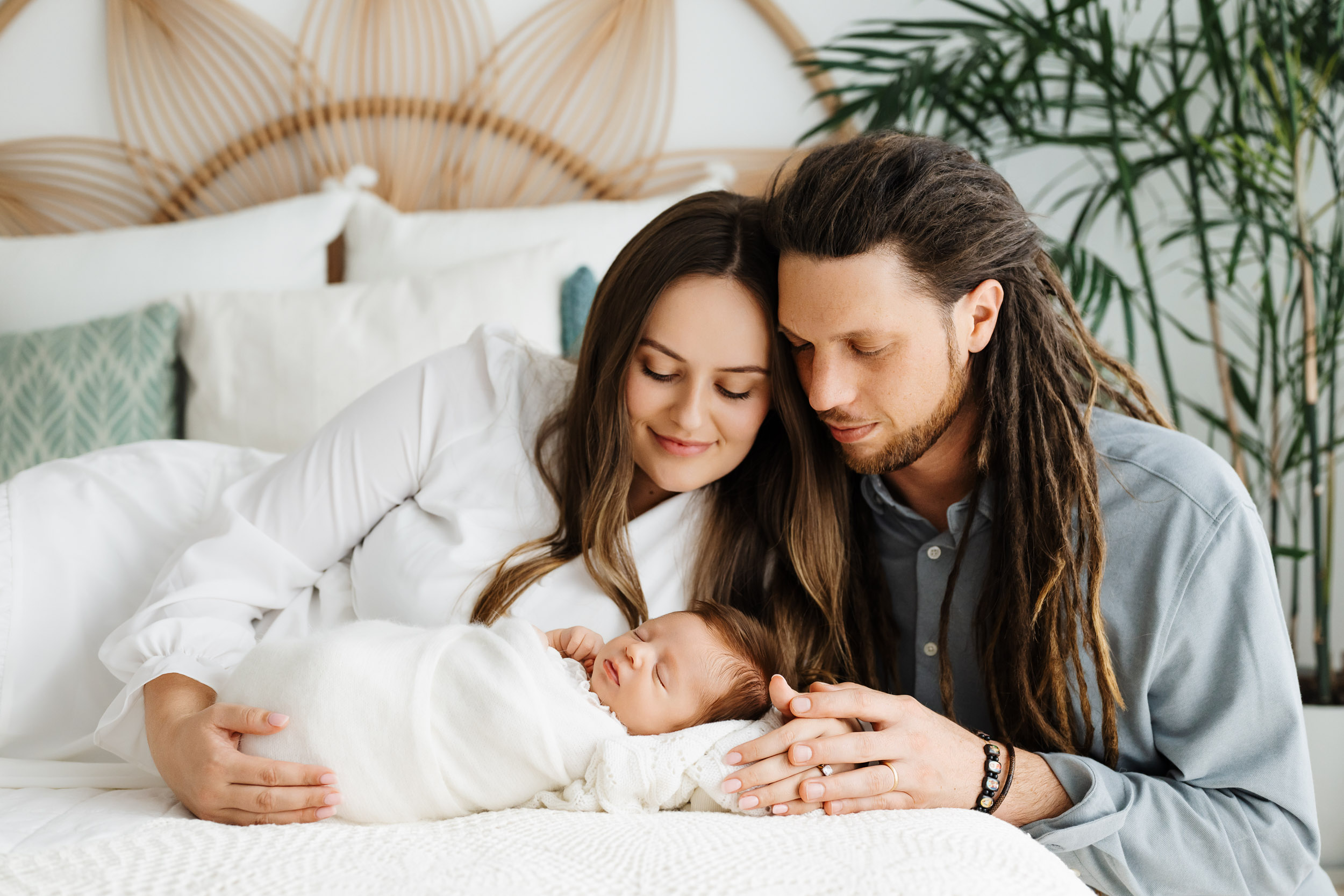 A couple with long hair gently touches a newborn baby wrapped in white on a bed, smiling softly.