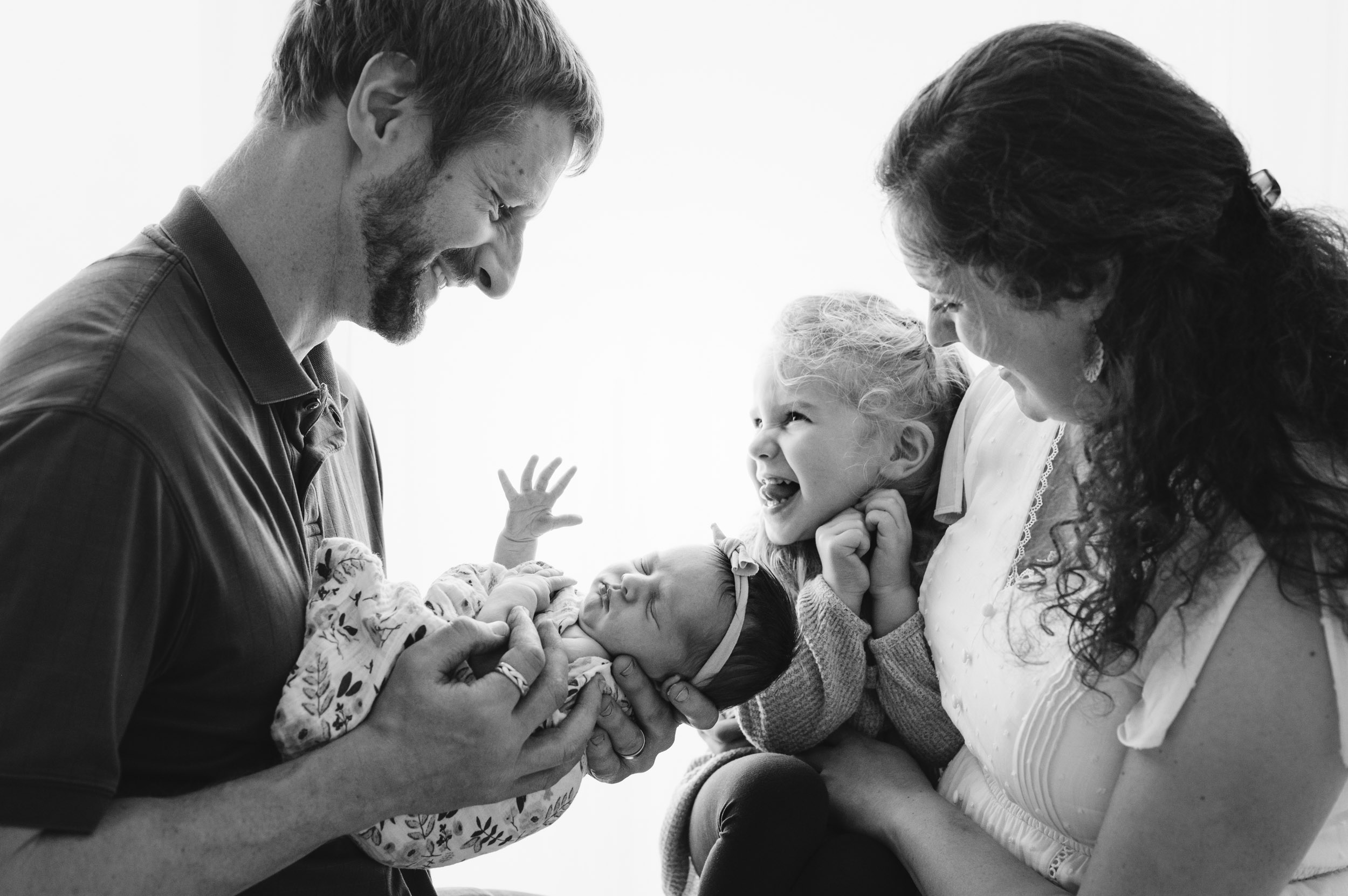 Family of four smiling and interacting with a newborn baby, with a young child touching the baby's hand, in black and white.