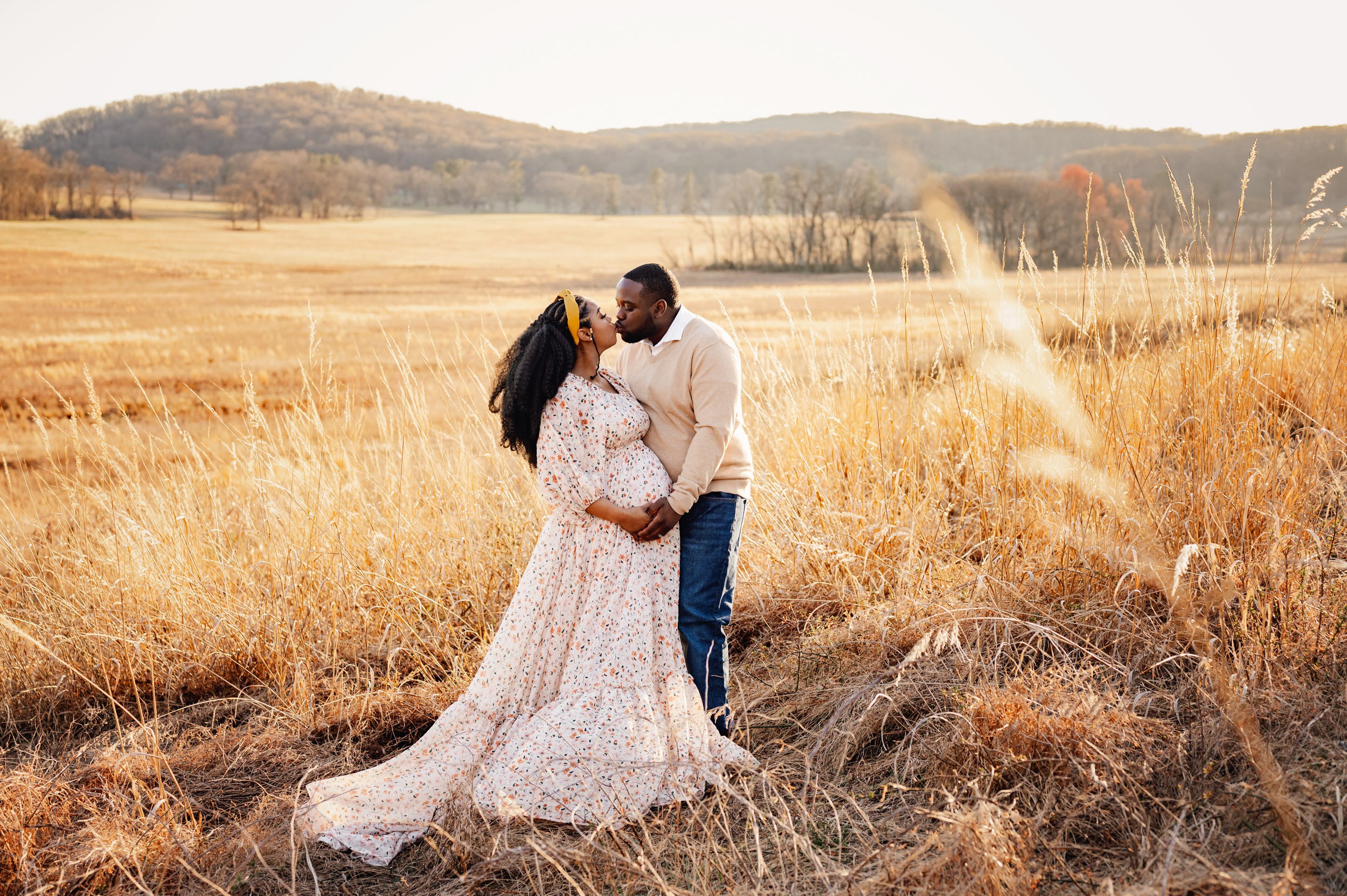 A couple stands close together in a field with tall grass, holding hands and looking at each other, with hills in the background.