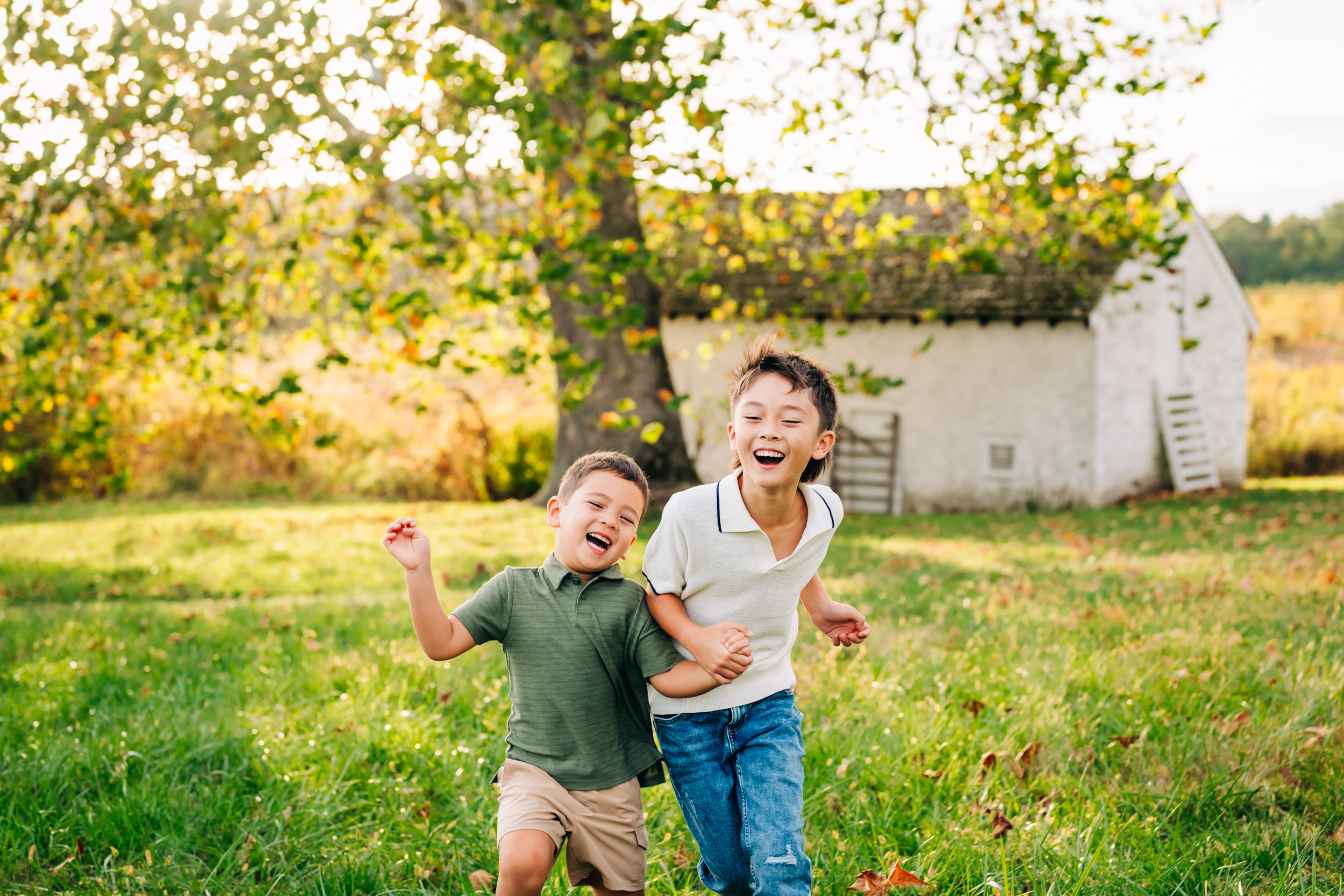 Two children laughing and playing outdoors in a grassy field with a tree and white building in background.