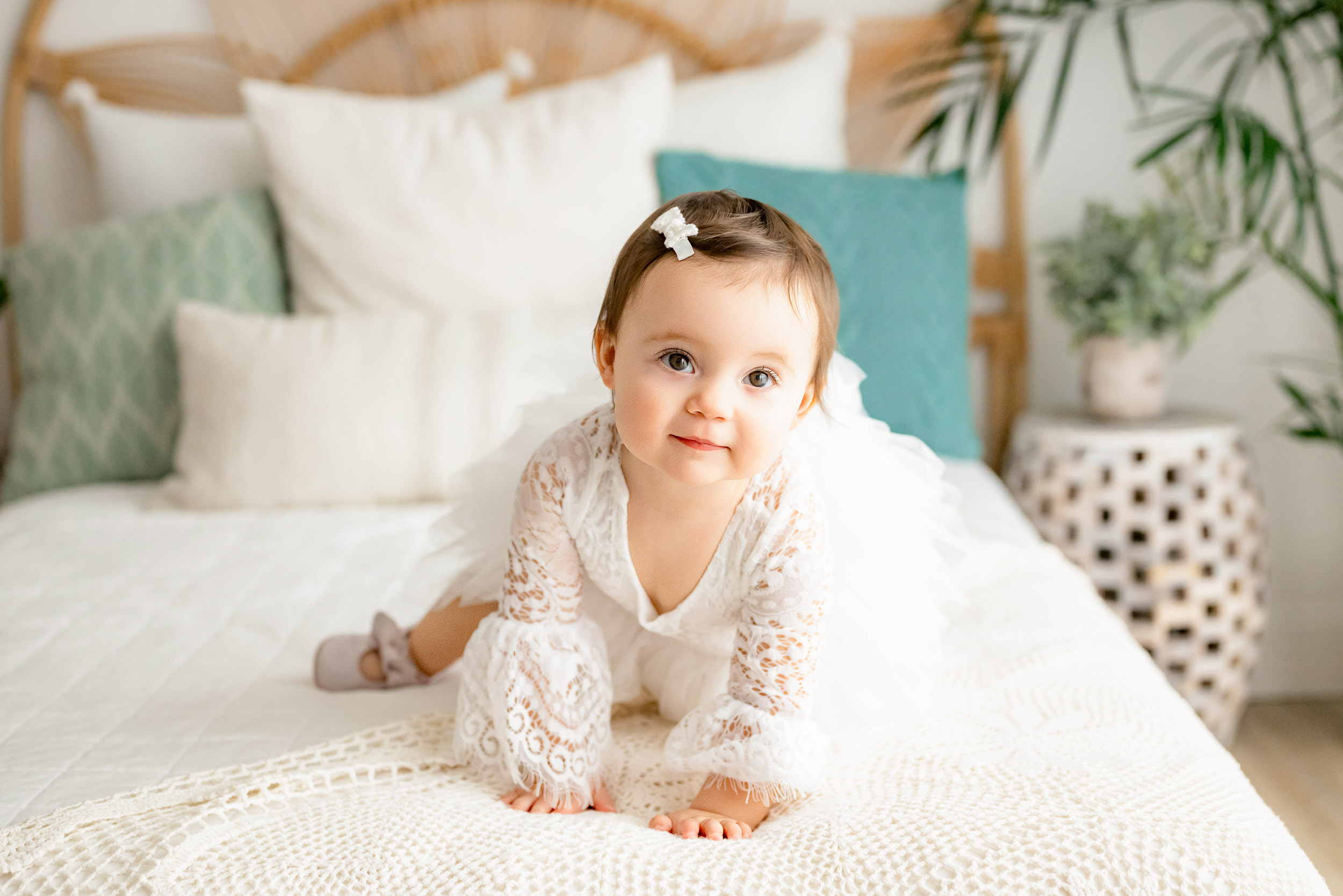 Baby girl crawling on bed with white lace dress and bow in hair, in a bright bedroom with pillows and plants