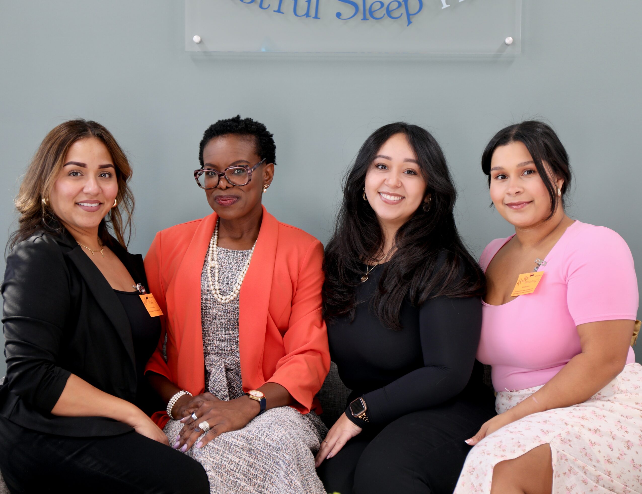 Four women sitting together in a room with a sign above them, smiling at the camera.