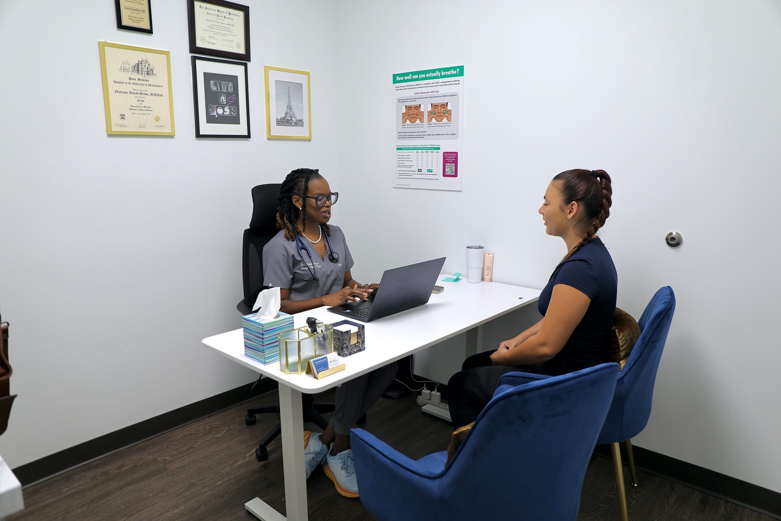 Two women sit across from each other at a white table in an office, engaging in conversation. One has a laptop, the other has a glass of water.
