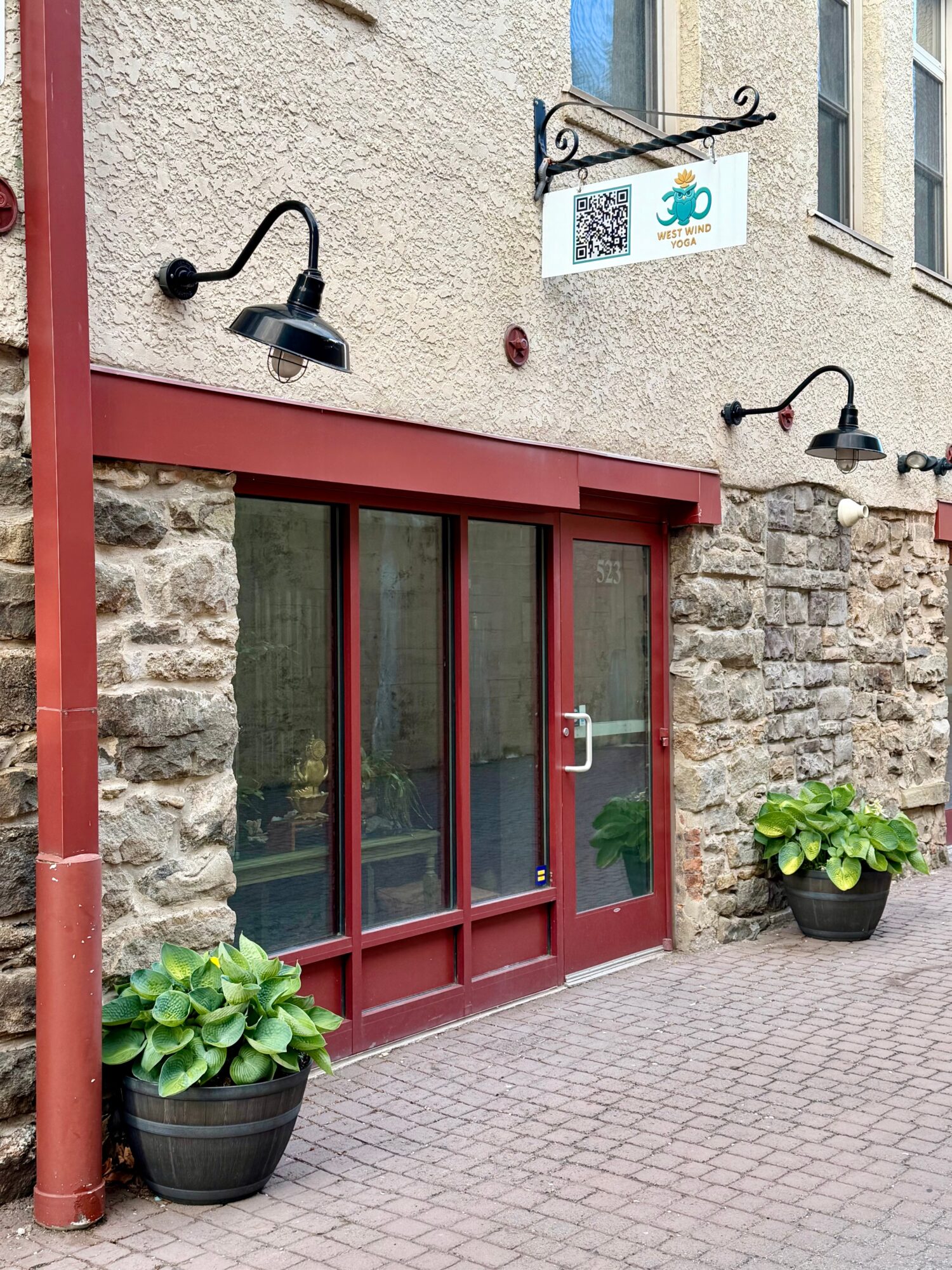 Exterior of a building with glass door, stone walls, potted plants, and a hanging sign, daytime.