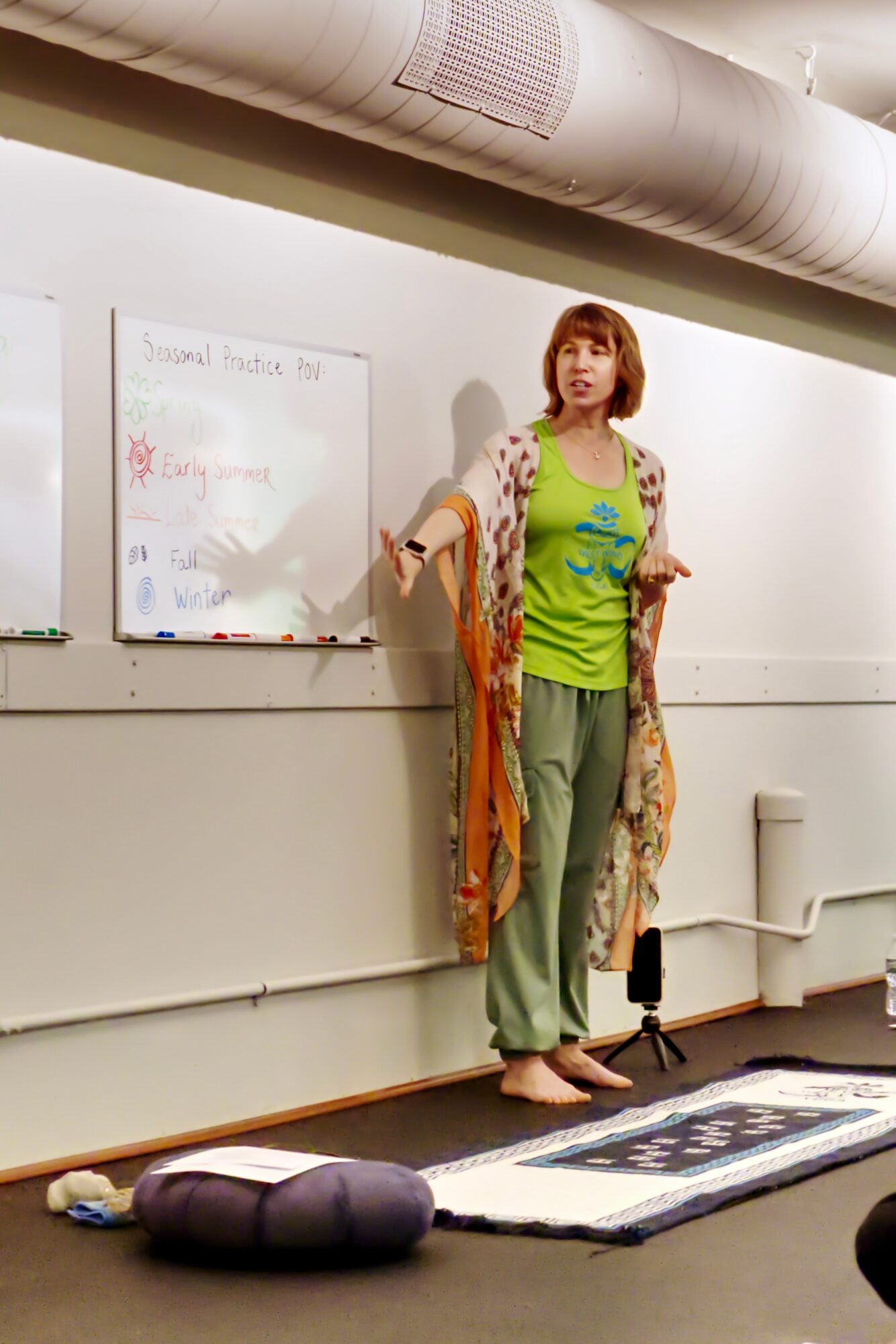 Woman standing in front of a whiteboard, holding a marker, with a colorful shawl, in a room with a white wall and ceiling pipe.
