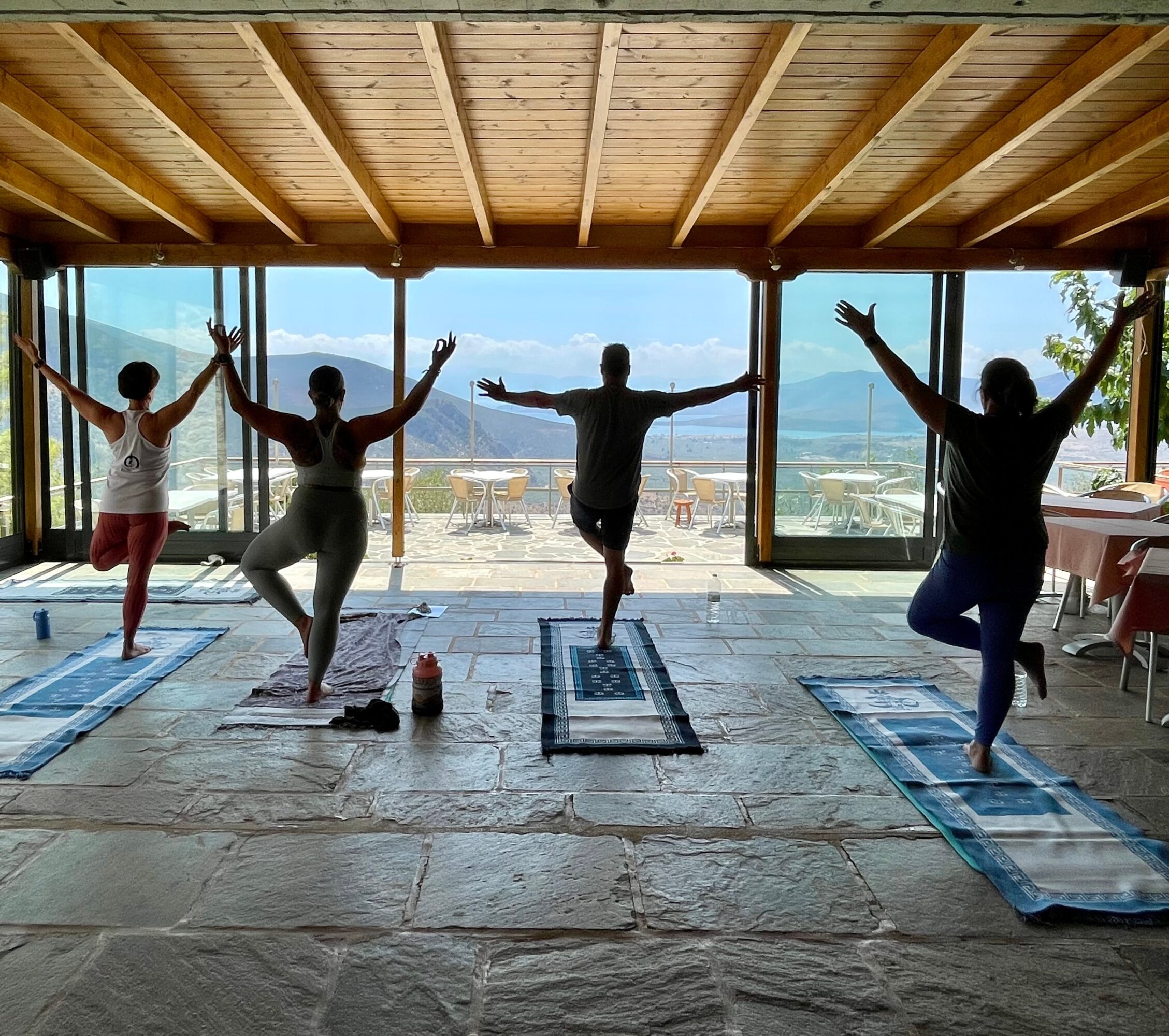 Four people practicing yoga indoors with large windows showing a scenic outdoor view, standing on mats with arms raised.