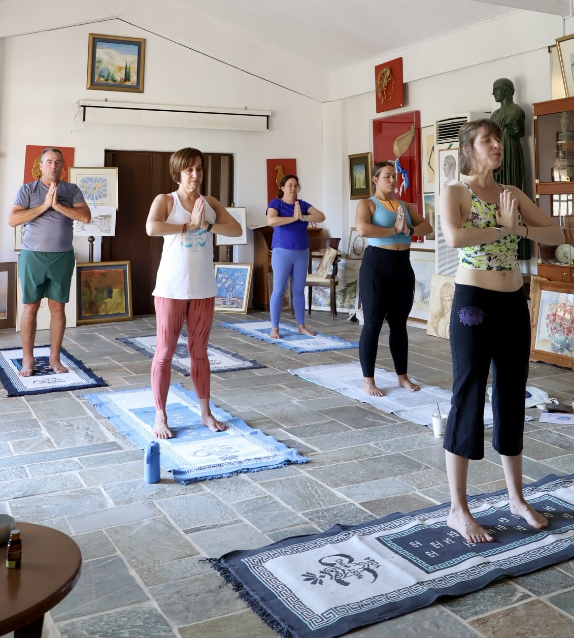 Group of people practicing yoga on mats in an art gallery with paintings and sculptures, standing with hands in prayer position.