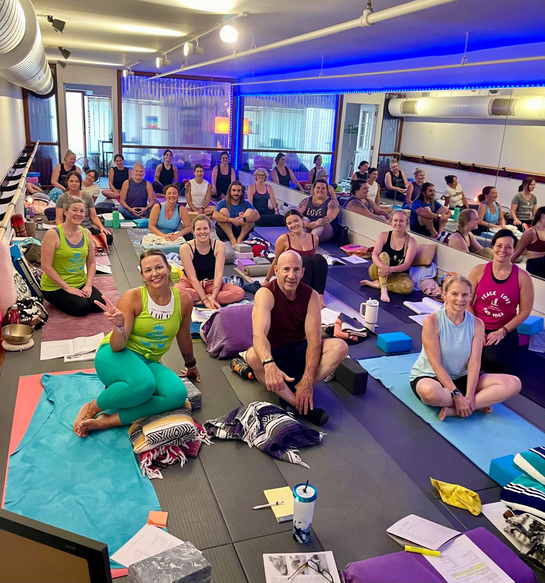 Group of people participating in a yoga class in a bright studio with large windows and blue lighting, sitting on mats.