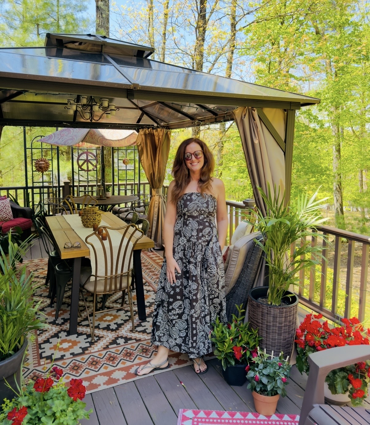 Woman in a floral dress standing on a porch with outdoor furniture and potted plants, surrounded by trees.
