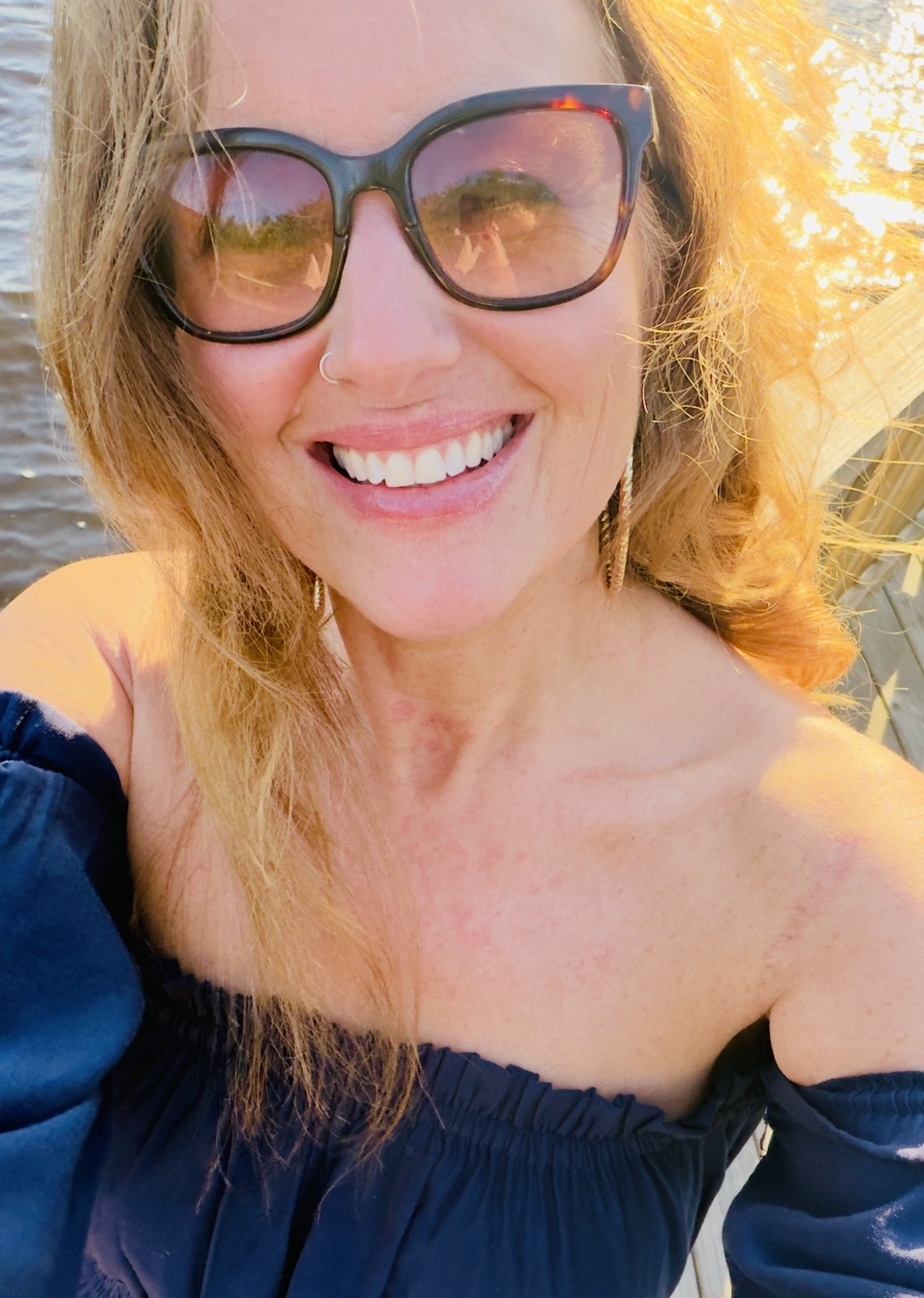 Smiling woman with glasses outdoors, sunlight, and yellow foliage in background.