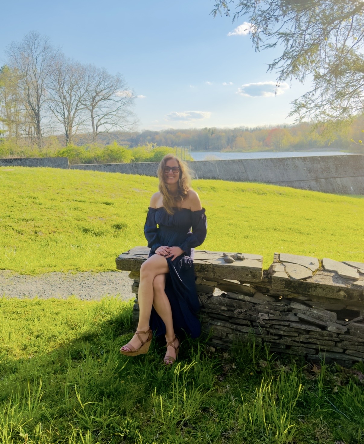 Woman sitting on a stone wall in a grassy park with trees and a river in the background, sunny day.