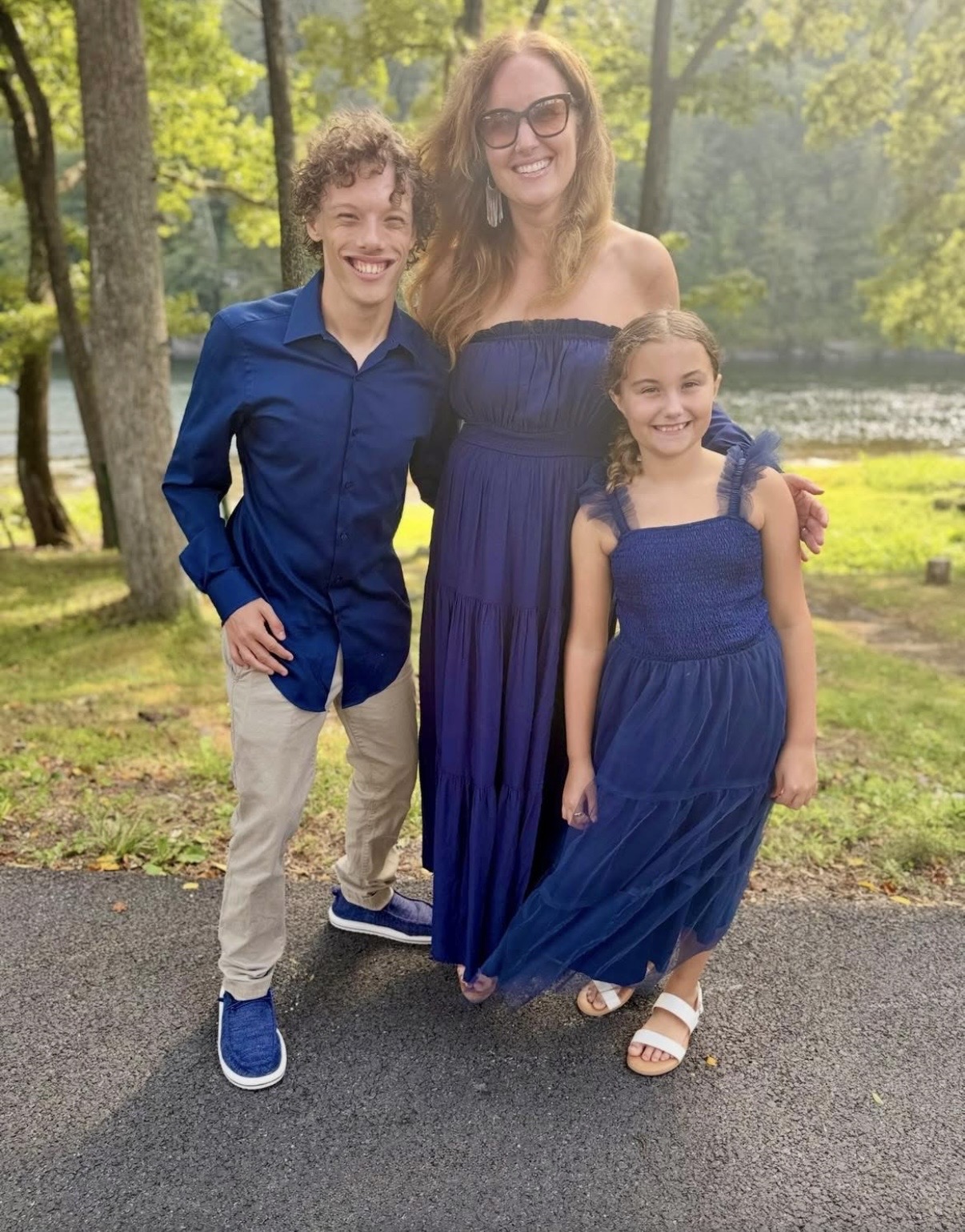 Three people, a woman and two children, standing outdoors on a path with trees and water in the background, smiling.