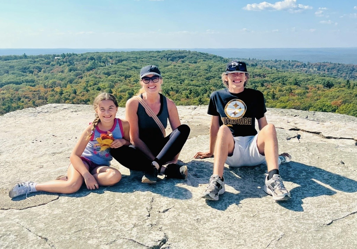 Three people sitting on a large rock with a scenic landscape and sky in the background.