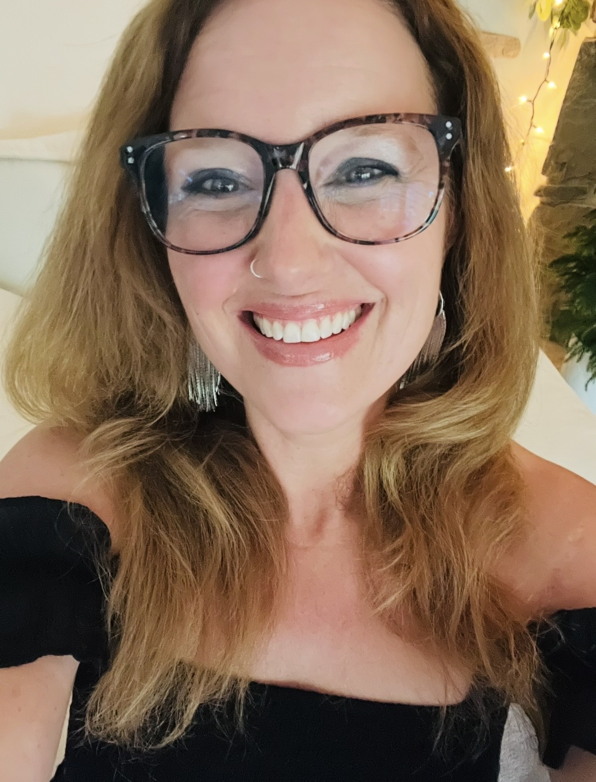 Smiling woman with glasses and wavy hair, wearing a black off-shoulder top, in a warmly lit indoor setting.