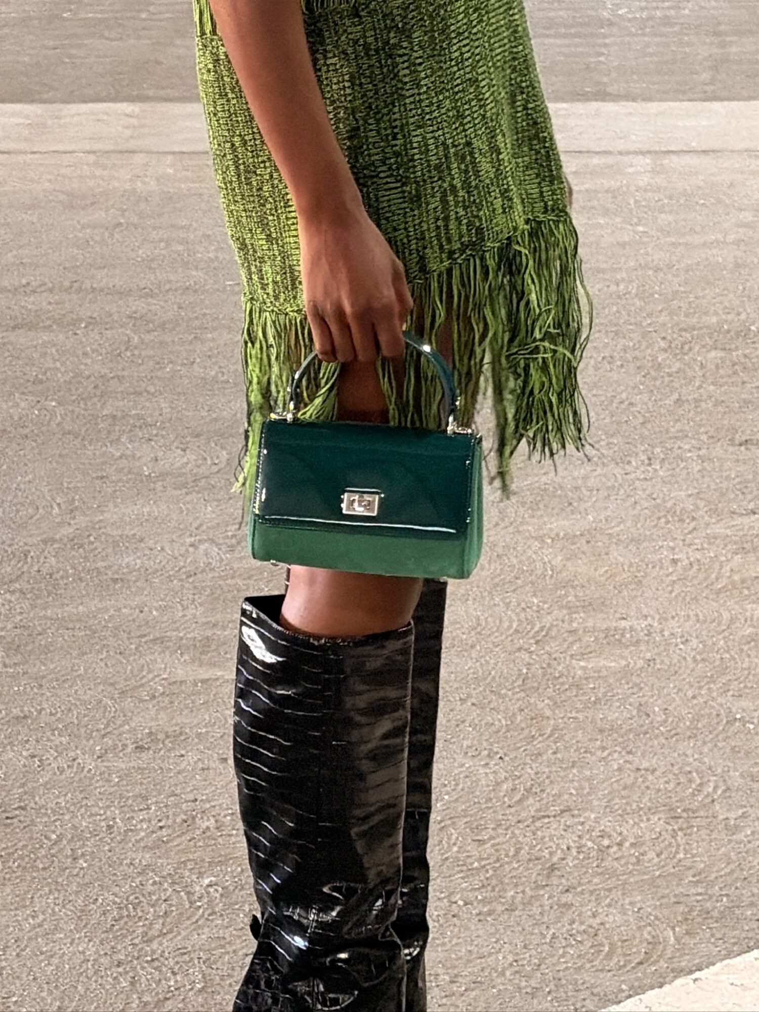 Person holding a small green handbag, wearing a green fringed dress and black shiny boots, standing on a concrete surface.