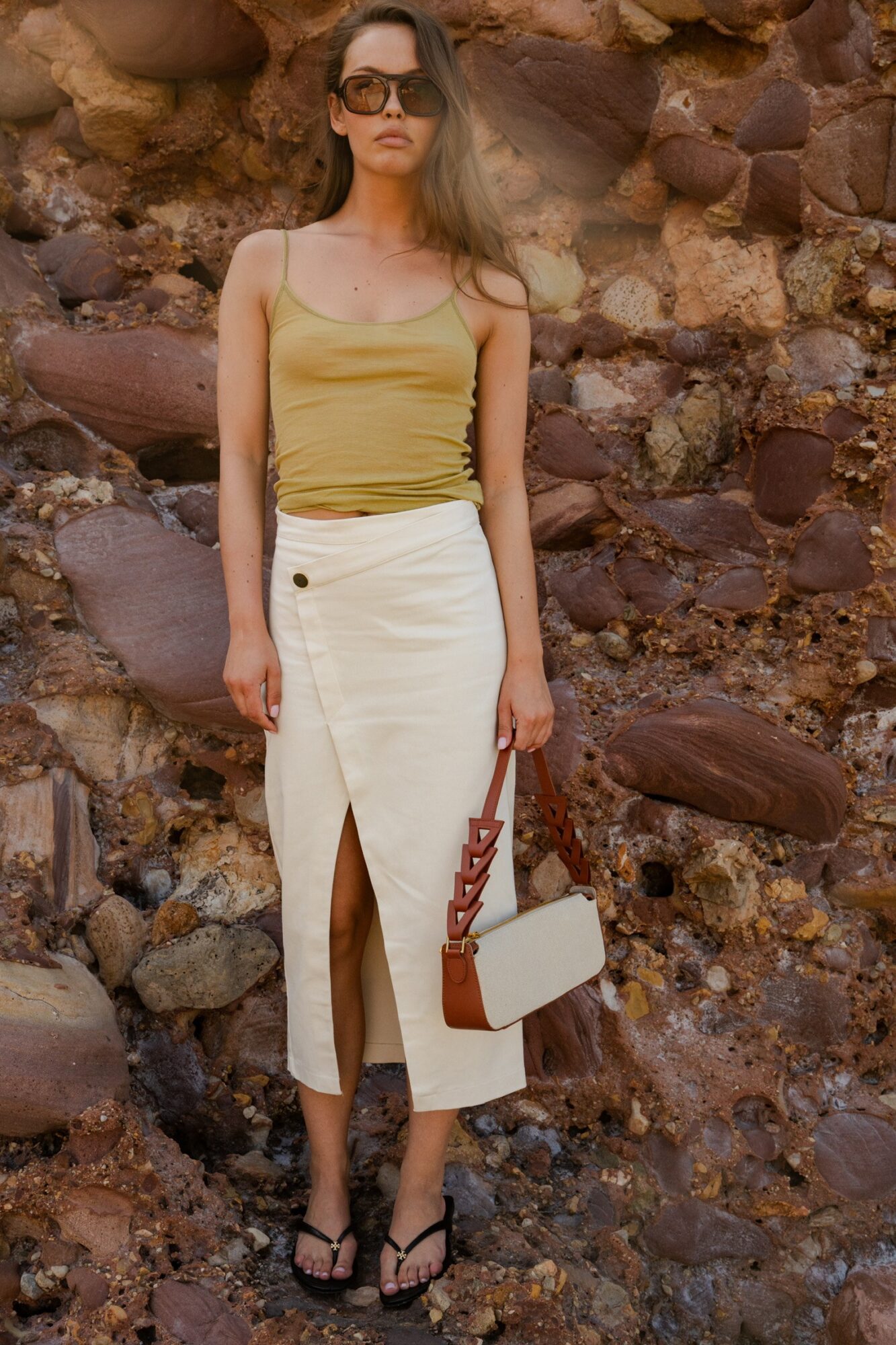 Woman in yellow top and white skirt standing against rocky background, holding a white bag with a brown strap.