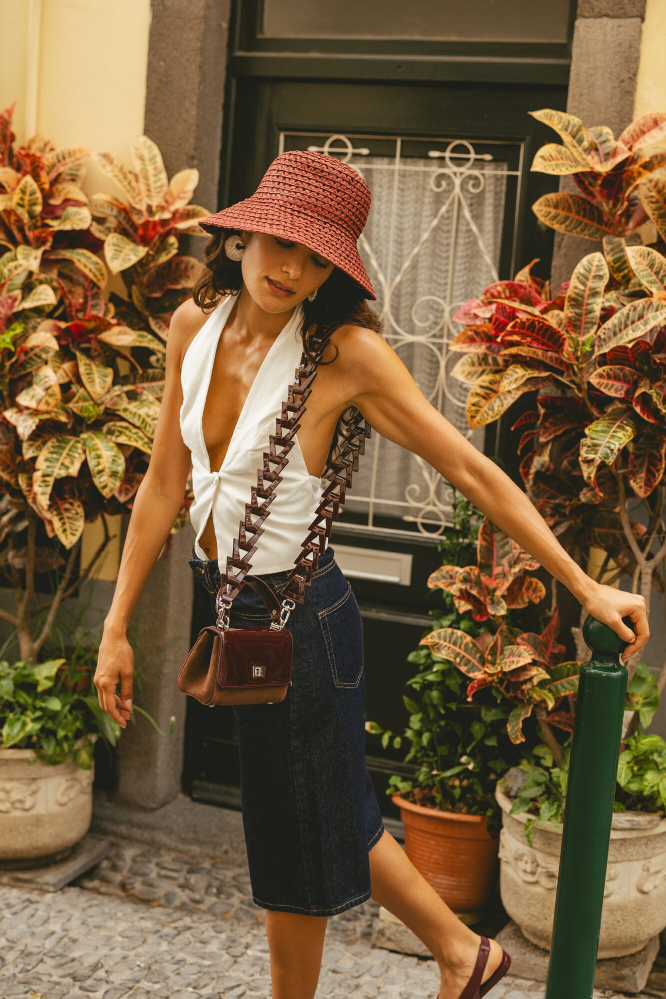 Woman in a pink hat, white top, and dark shorts standing among colorful plants outside a building.