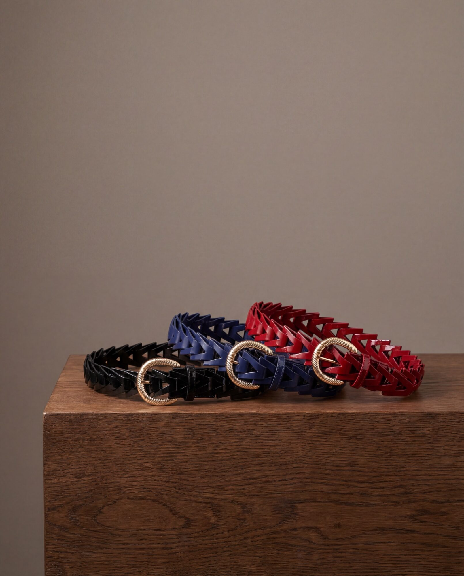 Three colorful belts with circular buckles are displayed on a wooden surface against a plain background.