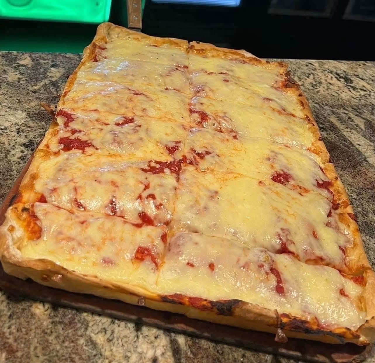 Large rectangular cheese pizza on a granite countertop with melted cheese and tomato sauce visible.