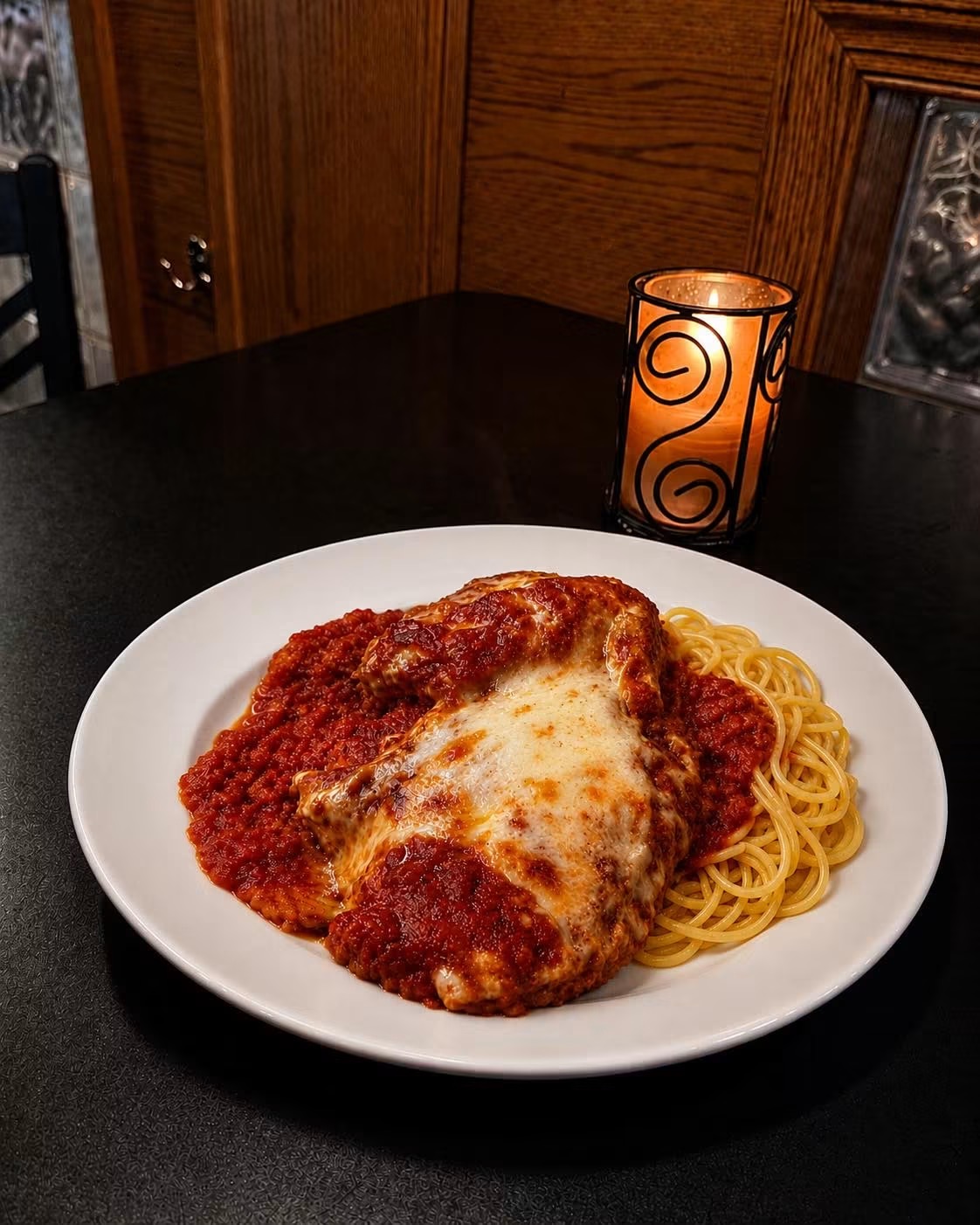 Plate of spaghetti with meat sauce and melted cheese, on a dark table with a lit candle and wooden wall in background.