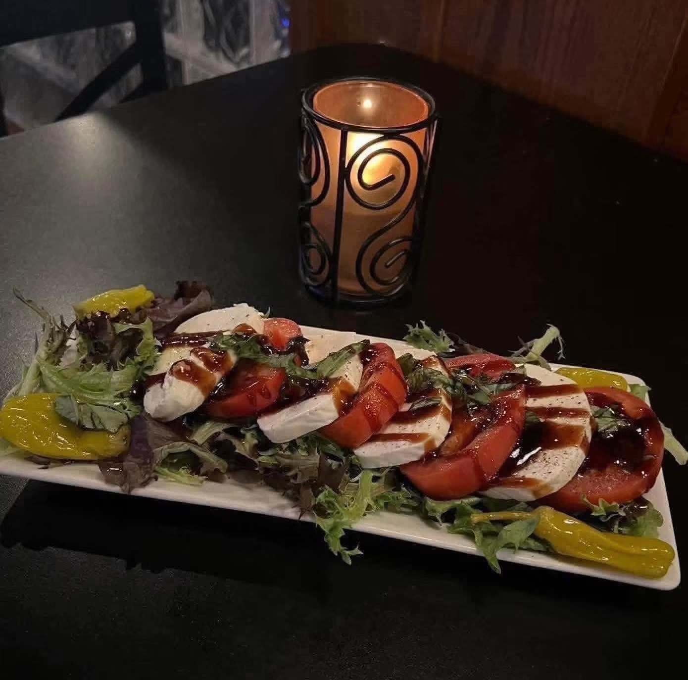 A long rectangular plate with sliced tomatoes, mozzarella, and basil, garnished with greens and yellow peppers, on a dark table with a lit candle.