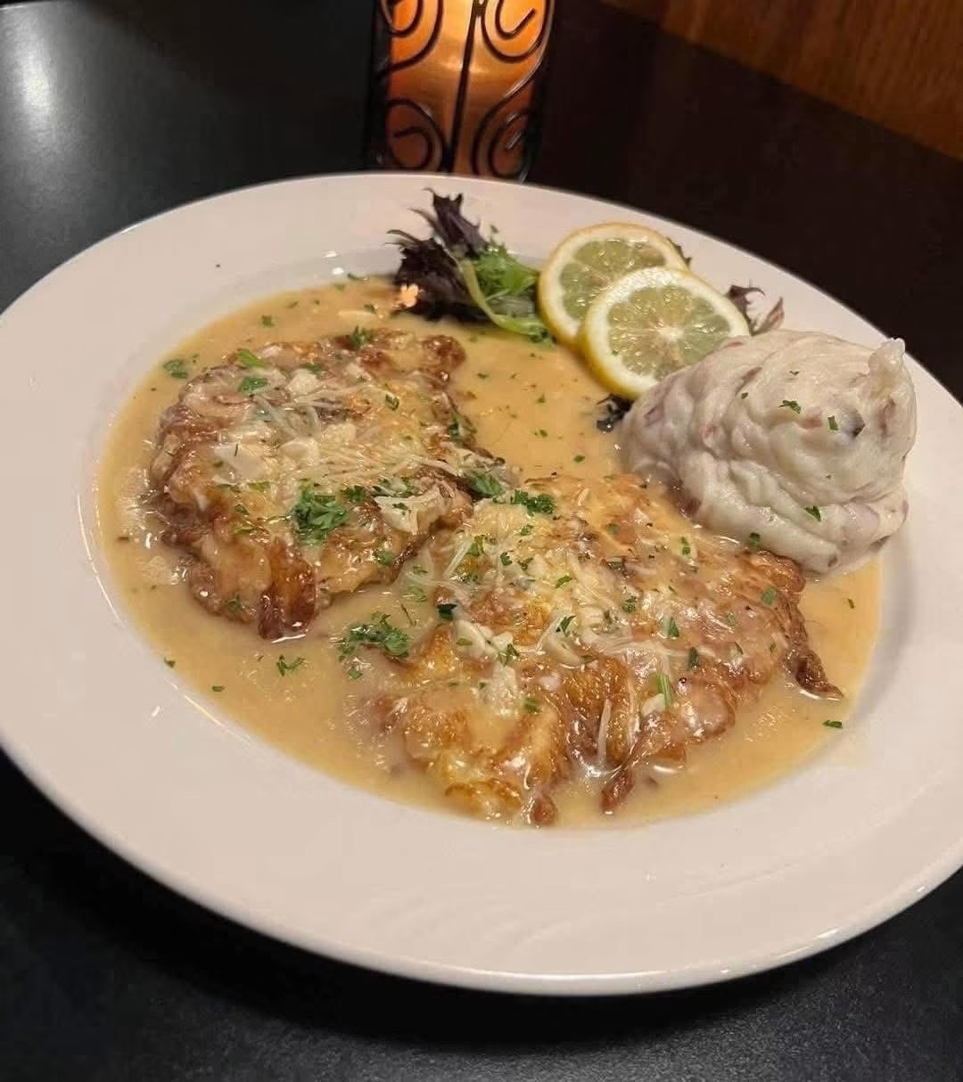 Plate with two pieces of fish in creamy sauce, lemon slices, and a dollop of mashed potatoes, garnished with herbs.