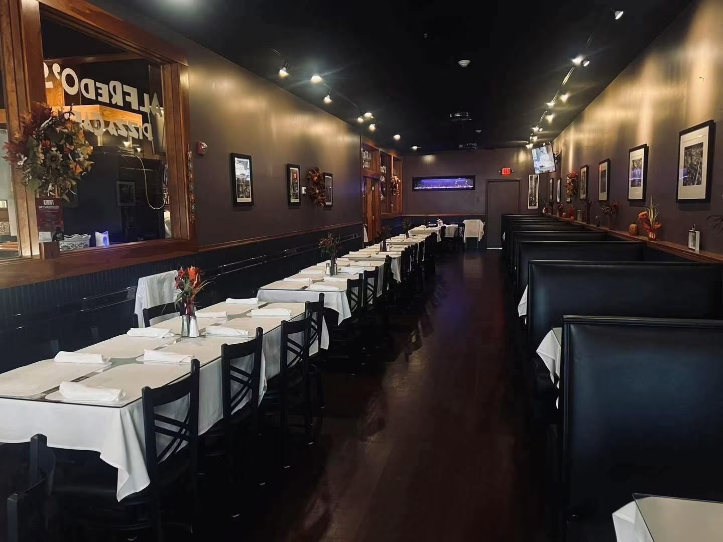 Long restaurant table with white tablecloths, black chairs, and framed pictures on dark walls, dim lighting, and a wooden floor.
