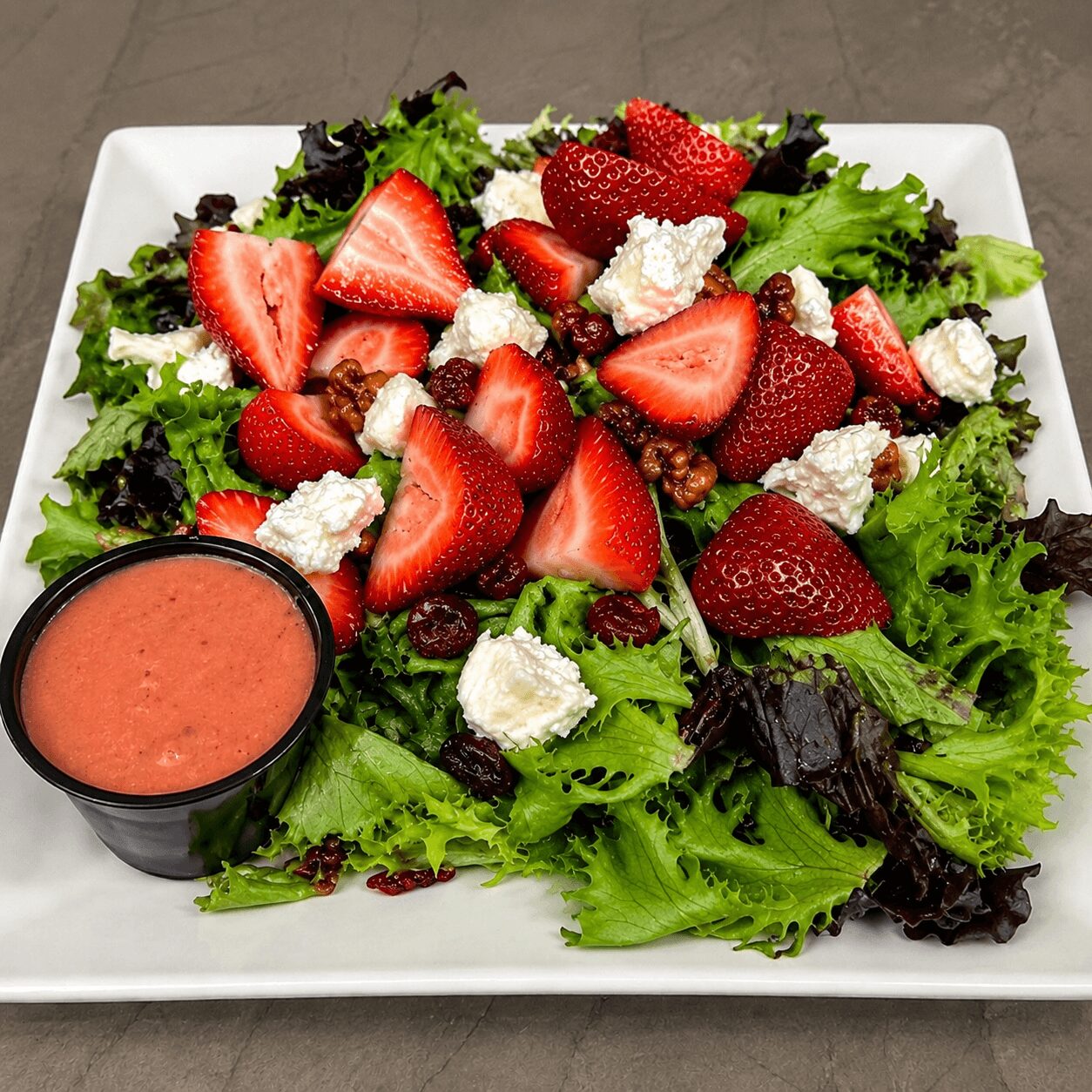 Mixed green salad with strawberries, crumbled cheese, and a side of pink dressing in a black cup on a white plate.