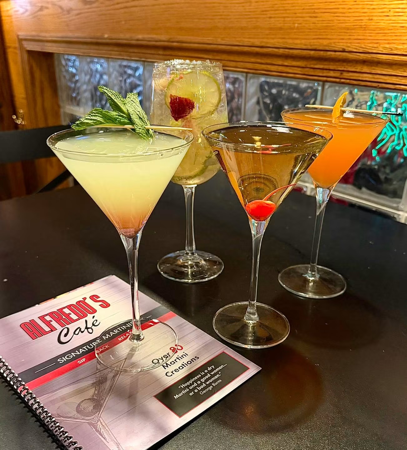 Four colorful cocktails on a table with a menu, in front of a window with a wooden frame.