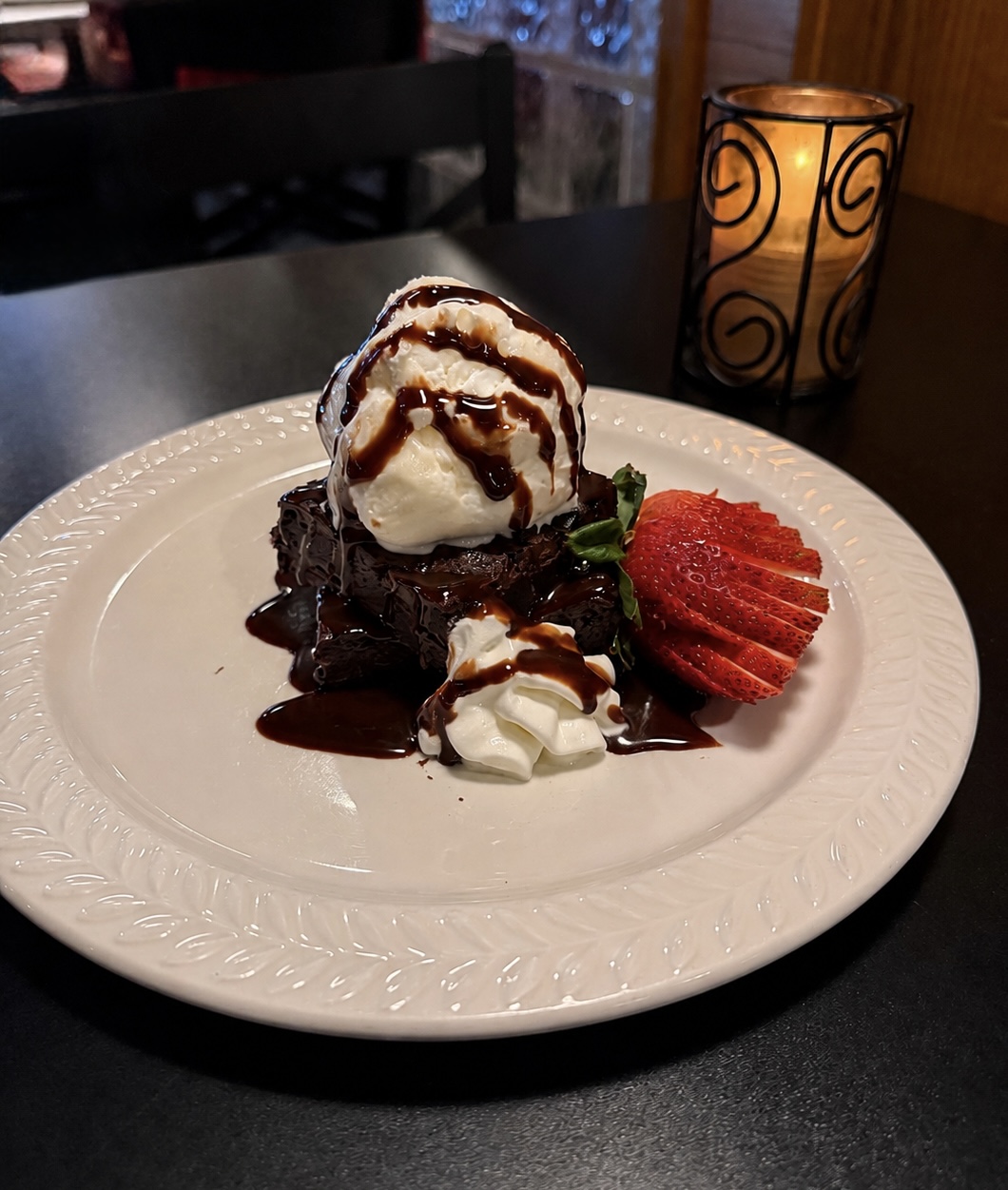 Dessert with ice cream, chocolate sauce, whipped cream, and sliced strawberries on a white plate, candle in background.