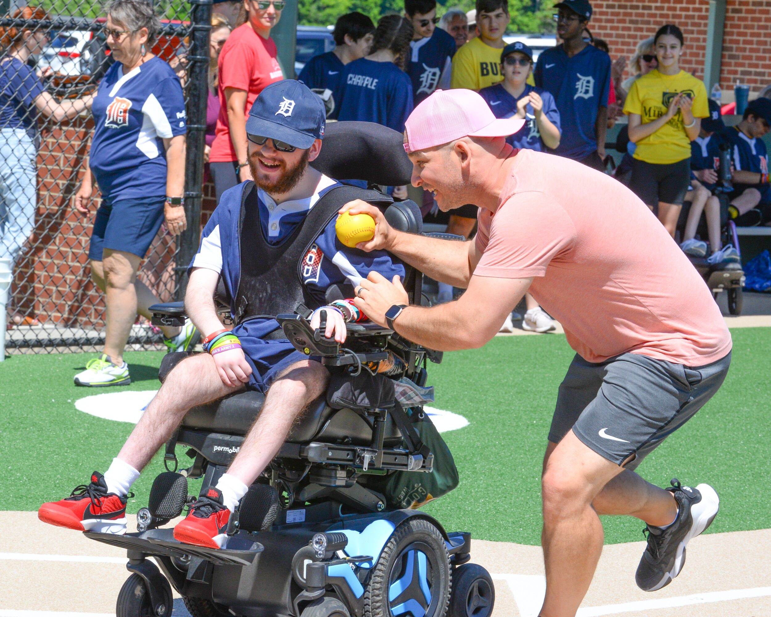 Person in a pink cap and pink shirt interacts with a person in a wheelchair holding a yellow ball, outdoors with people in the background.