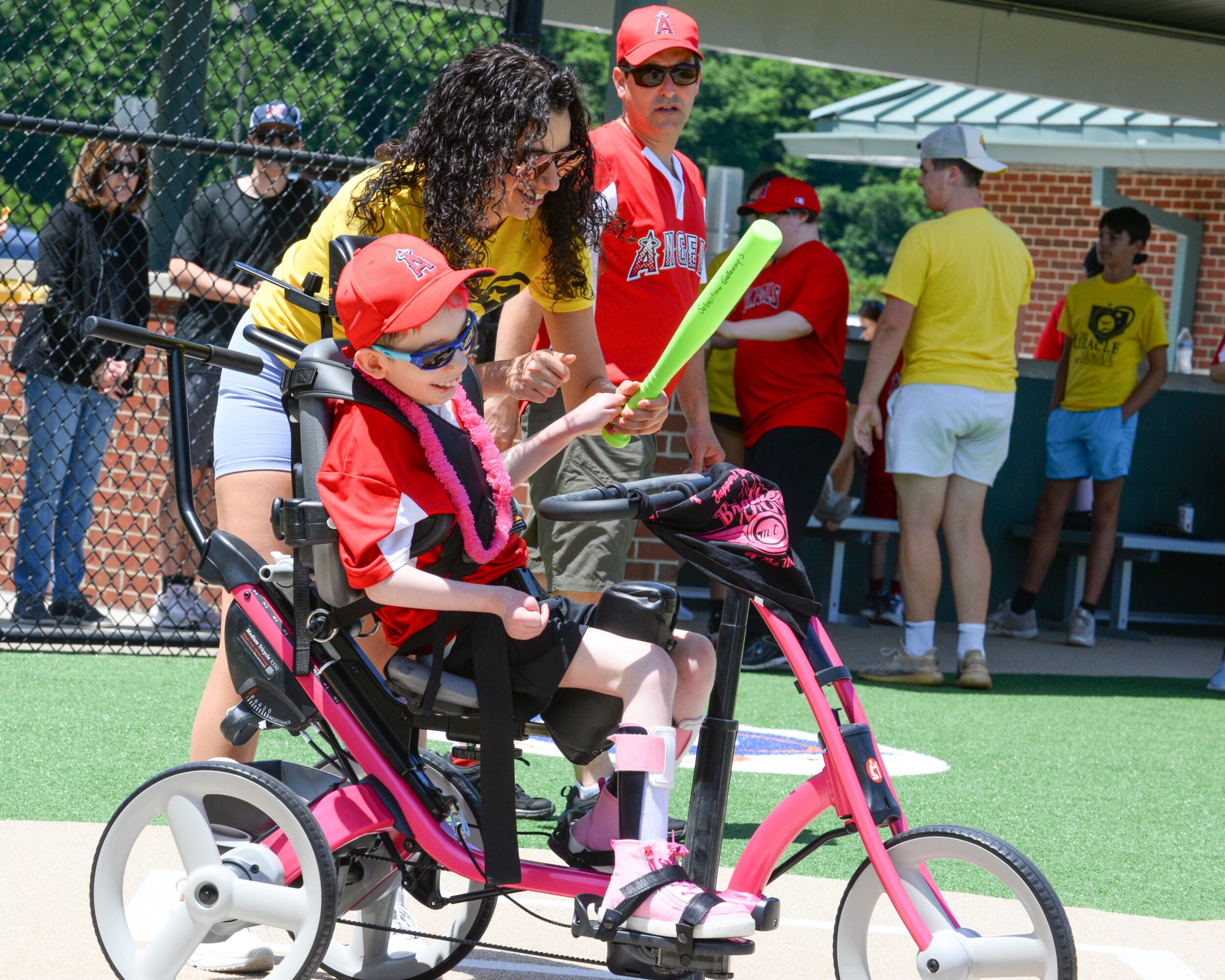 Child in a pink and black wheelchair holding a green balloon, with an adult woman beside her, outdoors at a sports field.