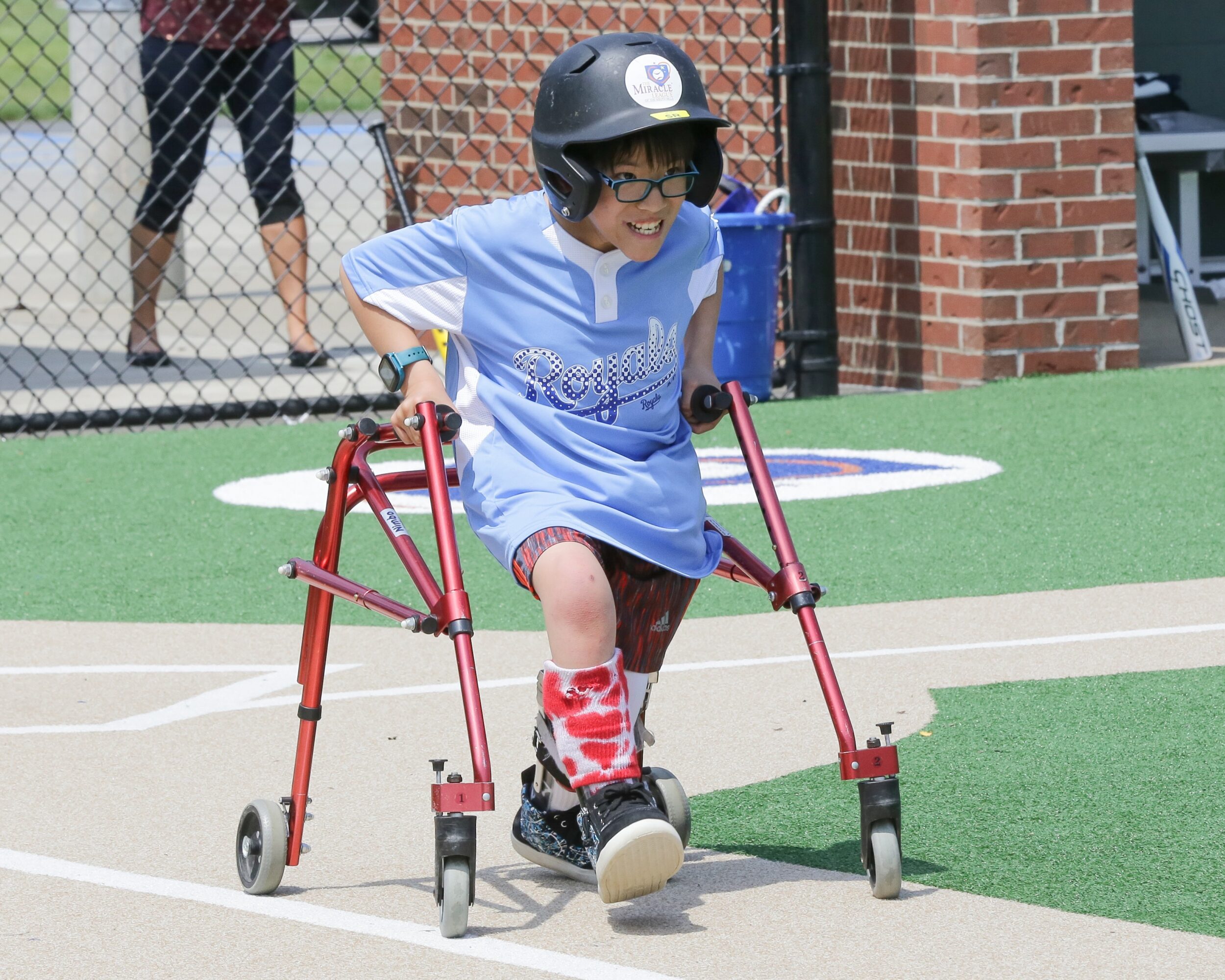 Child using a walker on a sports field, wearing a helmet and glasses, smiling, with a brick wall and fence in background.