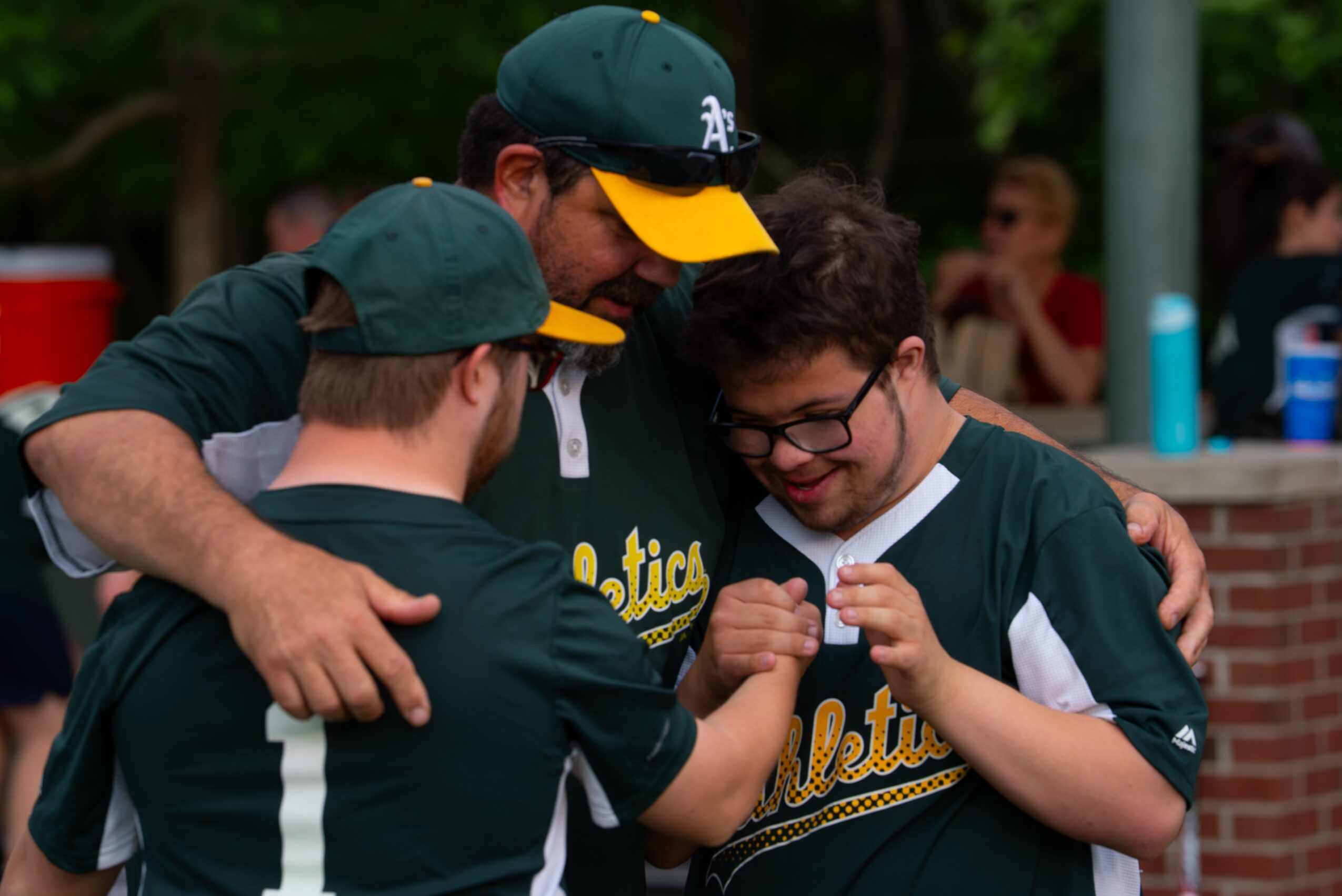 Three young people in baseball jerseys and caps embrace and smile together outdoors.