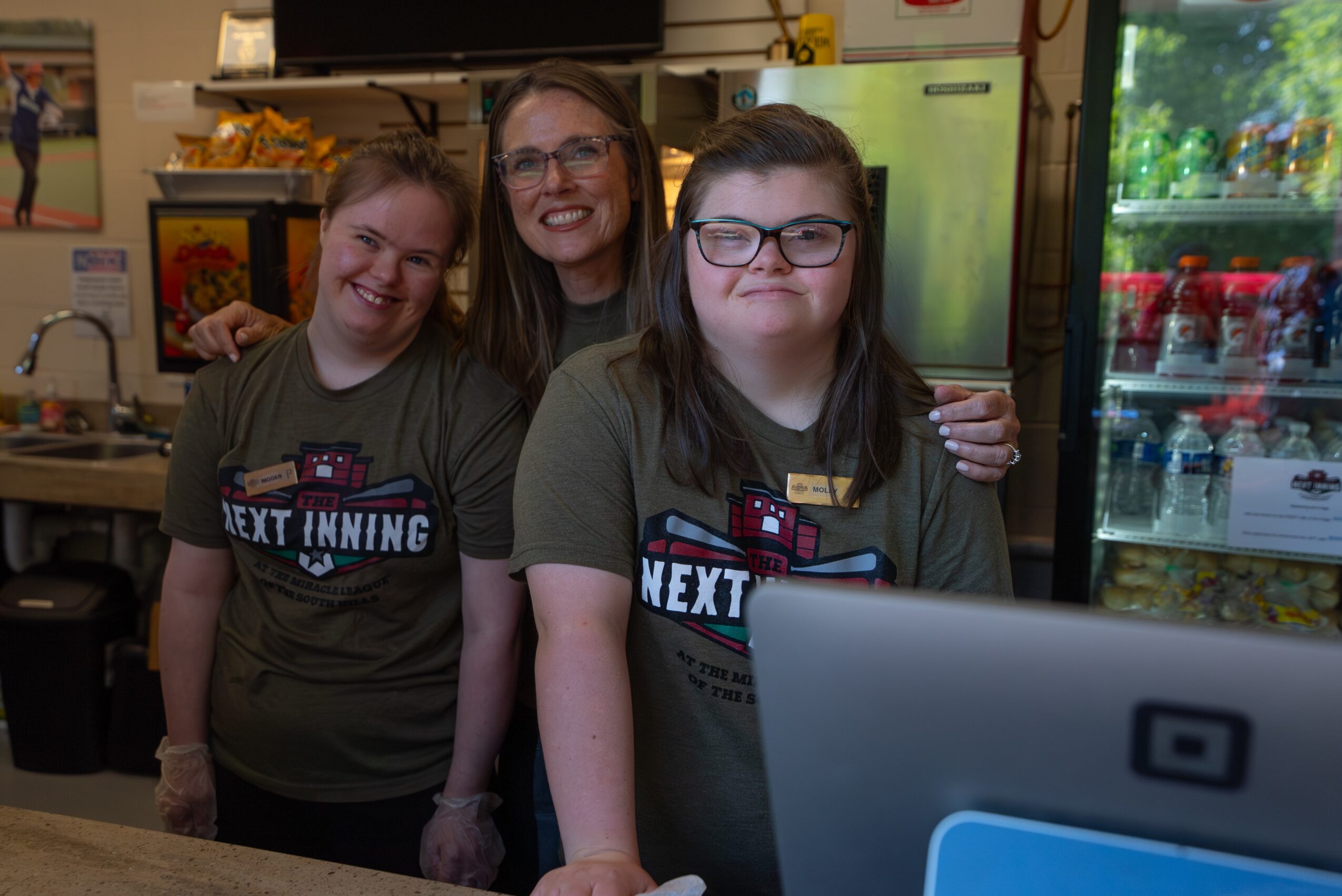Three smiling people standing in a kitchen, two young girls and an adult woman, with a refrigerator and a window in the background.