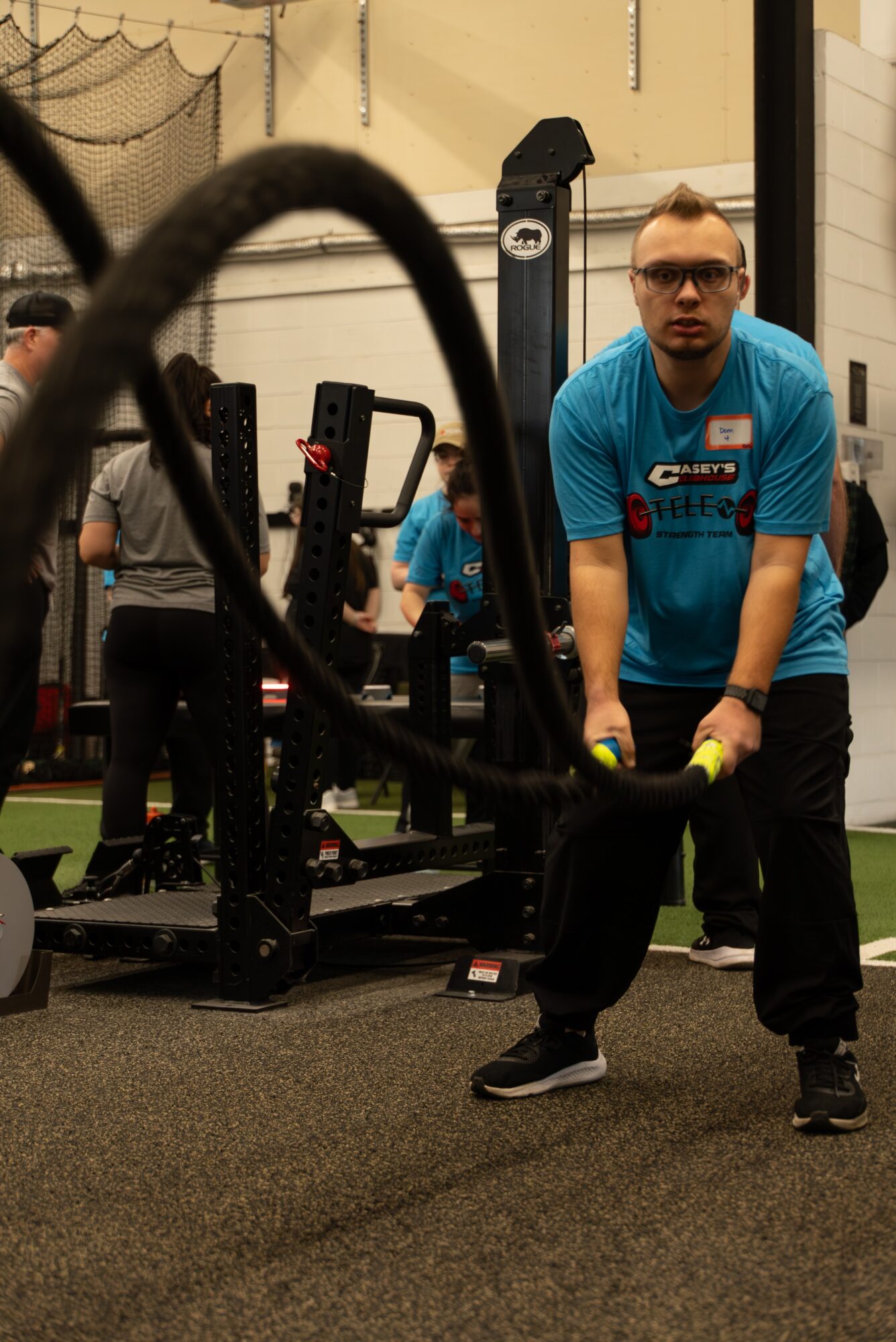 Person in blue shirt lifting a large tire during a fitness event indoors, with equipment and other people in background.