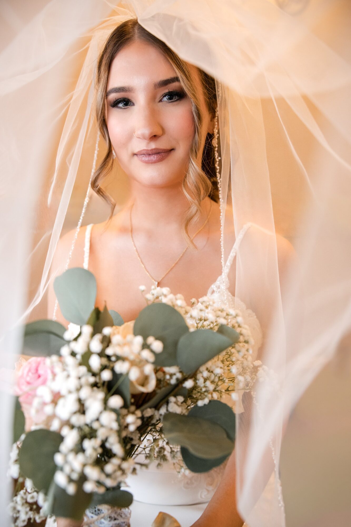 Young woman in wedding dress holding bouquet, smiling, with veil draped over her head, soft lighting, neutral background.
