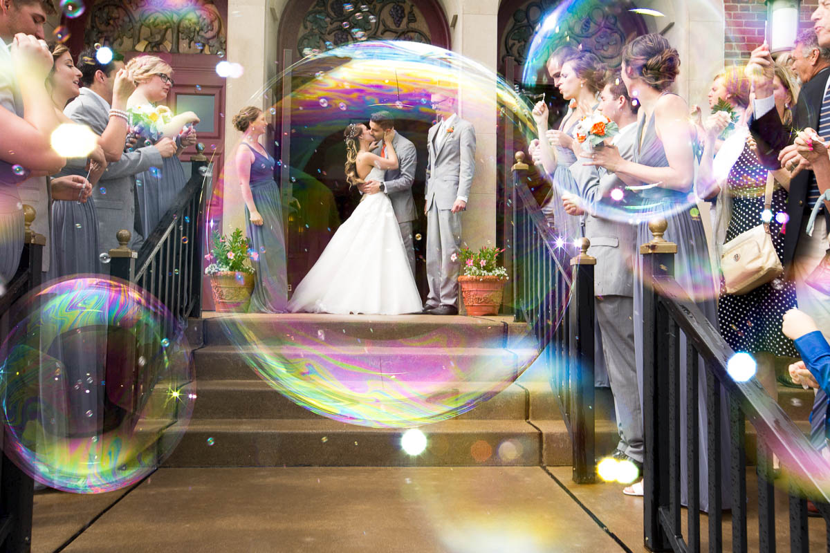 Wedding couple standing on stairs surrounded by guests, with large soap bubbles floating around them indoors.