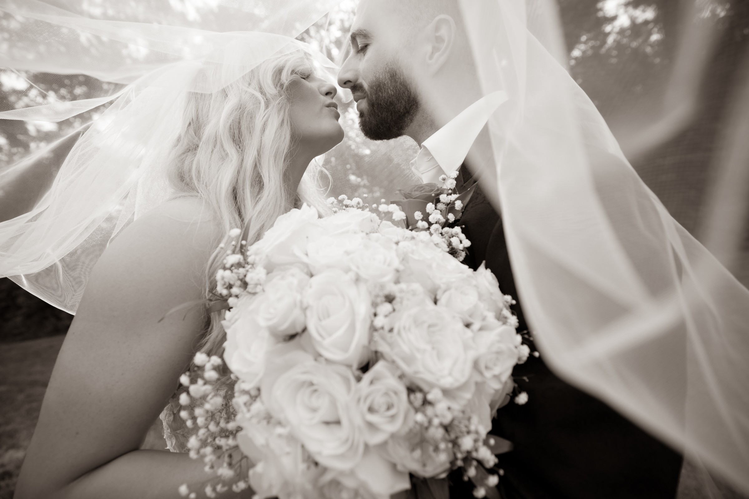 Couple with wedding veil and bouquet sharing a kiss outdoors, bride holding a large bouquet of roses, soft lighting.