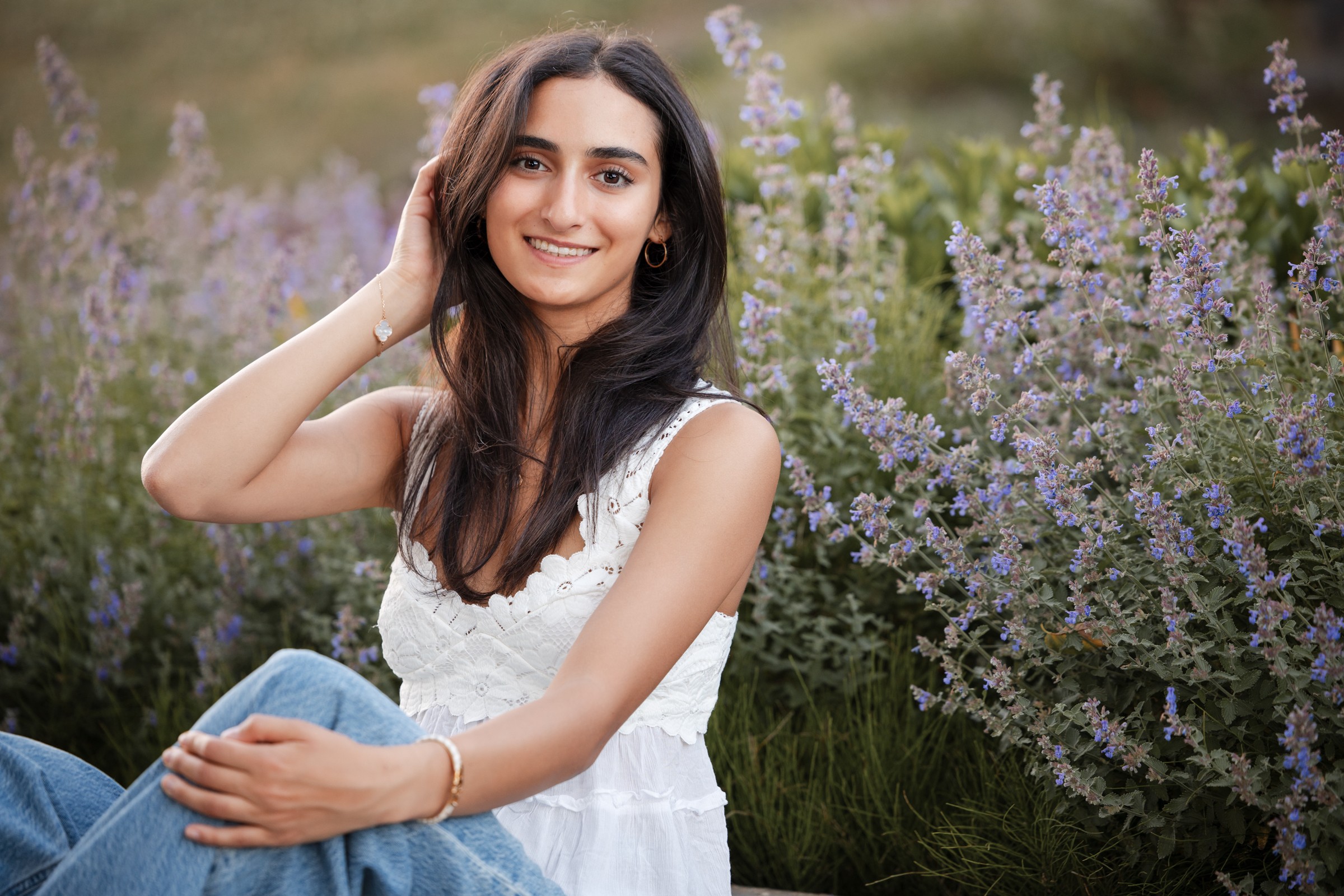 Young woman with long dark hair smiling outdoors among purple flowers, wearing a white sleeveless top and blue jeans.